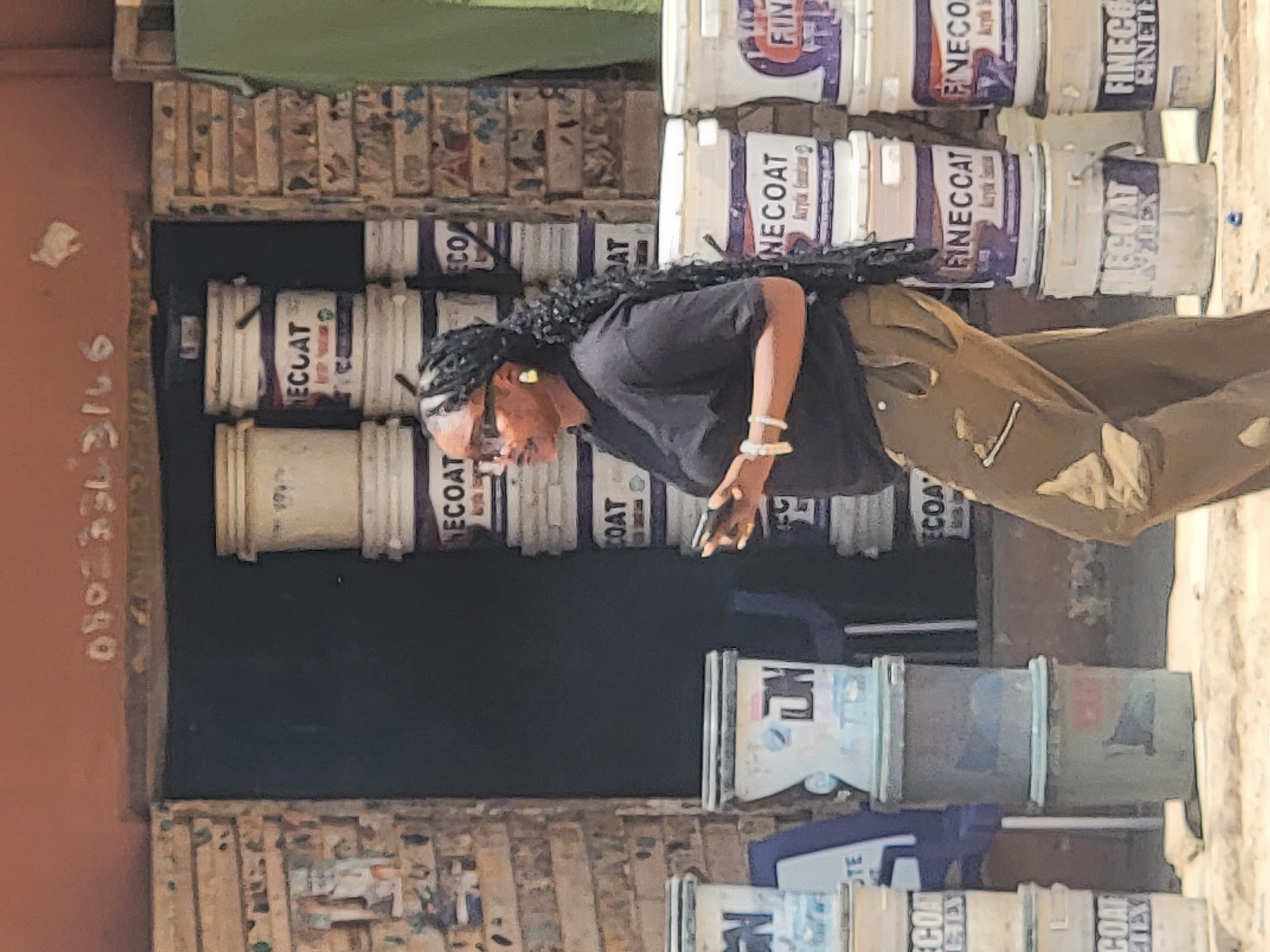 The scene captures a woman in Maiburiji, Nigeria, standing amidst a street setting with visible urban elements. She has dark curly hair and wears sunglasses and a black top paired with khaki pants. Her left hand is visible, and she seems to be holding a phone.
Behind her and to her left, there is a display of stacked paint buckets, predominantly white with purple lettering, against a darker backdrop. The text "FINECOAT Acrylic Emulsion" is visible on some of the buckets.
To the left of the frame is a reddish-brown wall with white writing. The foreground to the right is a sandy colored, slightly blurred area. Overhead, there's a glimpse of a green sheet or cloth. The overall impression is a snapshot of everyday life in an urban commercial environment in Maiburiji.