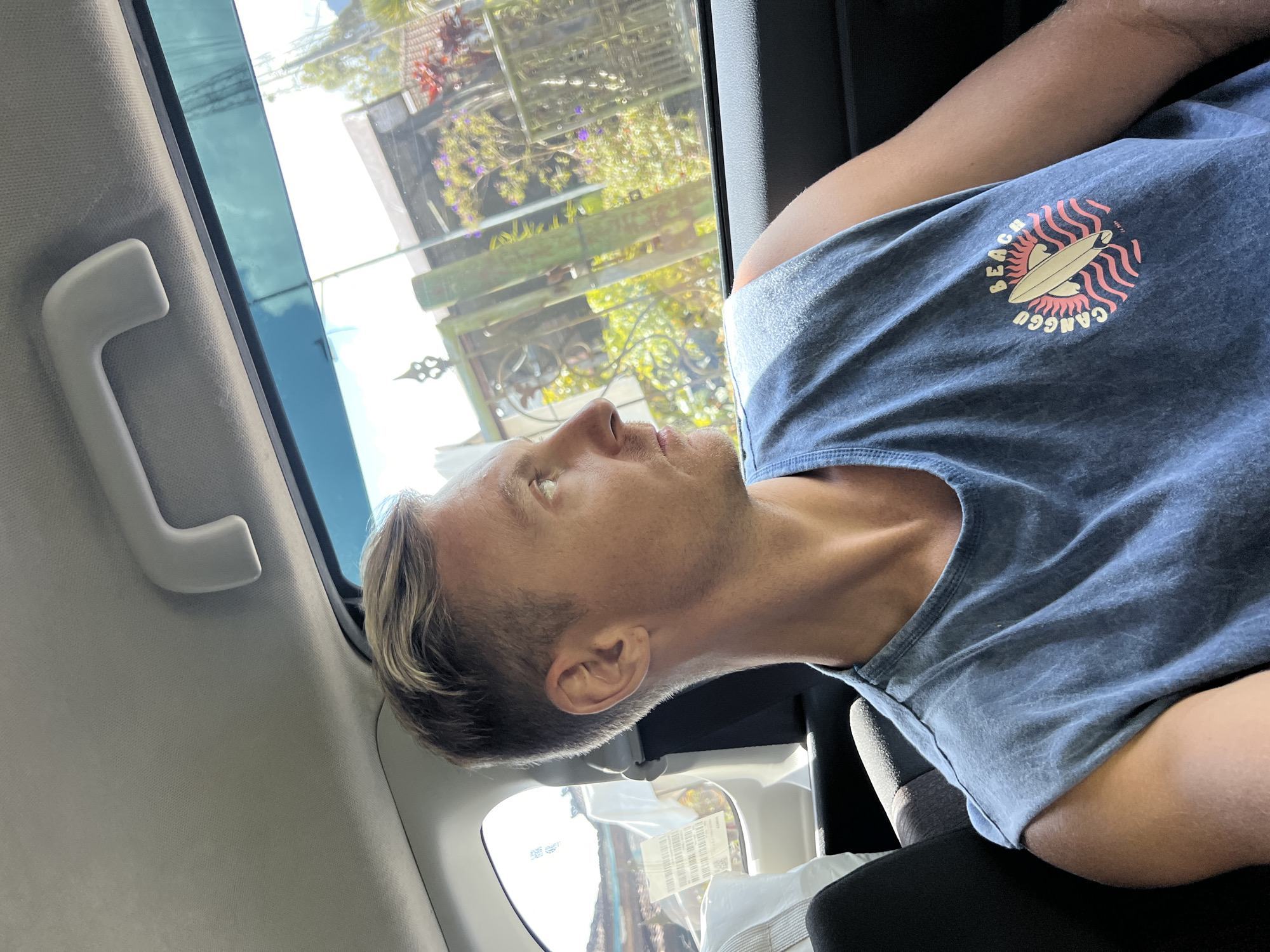 The image shows a person sitting in a car, possibly in Baturiti, Indonesia, judging from the writing on the person's shirt and the outdoor scenery. The person has light skin, blond hair, and is wearing a blue t-shirt with a graphic design that includes the words "Beach Canggu". The environment is a car interior. Outside the car window, there is a view of a garden or roadside greenery. The person is looking out the window.