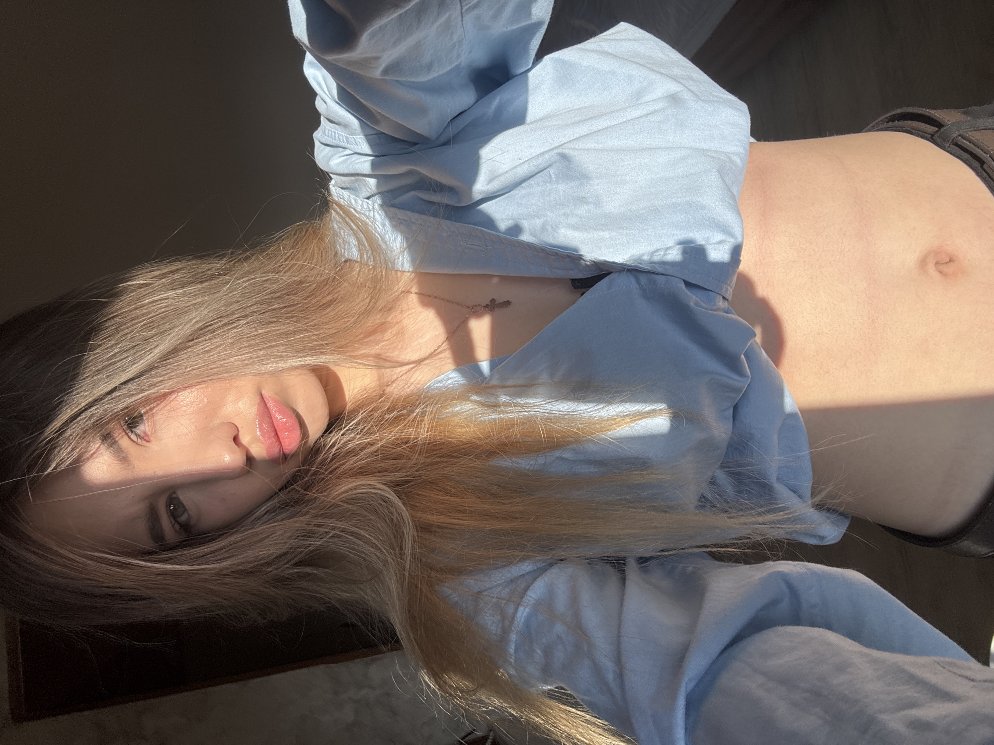 The image, possibly captured in Noviy, Russia, features a young woman lying down indoors, illuminated by strong, natural sunlight. Her head is positioned towards the left of the frame, with her body extending to the right. She has long, flowing light brown hair, which is partially sun-drenched and frames her face. Her eyes are visible, and her lips appear glossy.
She is wearing a light blue, long-sleeved shirt, styled to reveal her bare midriff. A delicate silver necklace with a small cross pendant is visible on her chest. Dark-colored clothing, possibly pants or shorts, with a visible belt, can be seen around her lower torso.
The intense, direct sunlight creates dramatic contrasts of light and shadow across the scene. The left portion of the image is largely in deep shadow, while the right side, encompassing much of her body and hair, is brightly lit, accentuating textures and details. The woman maintains a neutral or serene expression while posing for the photograph. The setting appears to be an interior space, likely a room or on a surface like a bed or floor, given the indoor lighting and background. There is no visible text in the image.