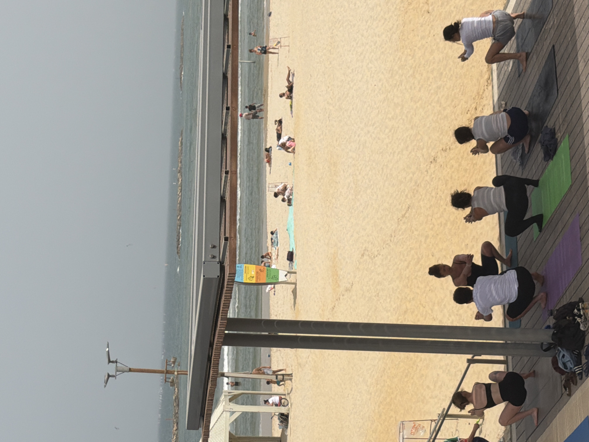 The image captures a scene at a beach in Tel-Aviv, Occupied Palestine. The environment consists of a sandy beach, the sea, and a partly cloudy sky. People are engaging in various activities. Near the shoreline, individuals are sunbathing or relaxing on the sand. On a boardwalk adjacent to the beach, a group is participating in a yoga class, using yoga mats. There are several objects, including beach umbrellas, towels, and personal belongings scattered on the sand. The image is taken from an elevated perspective, offering a broad view of the beach and surrounding area.