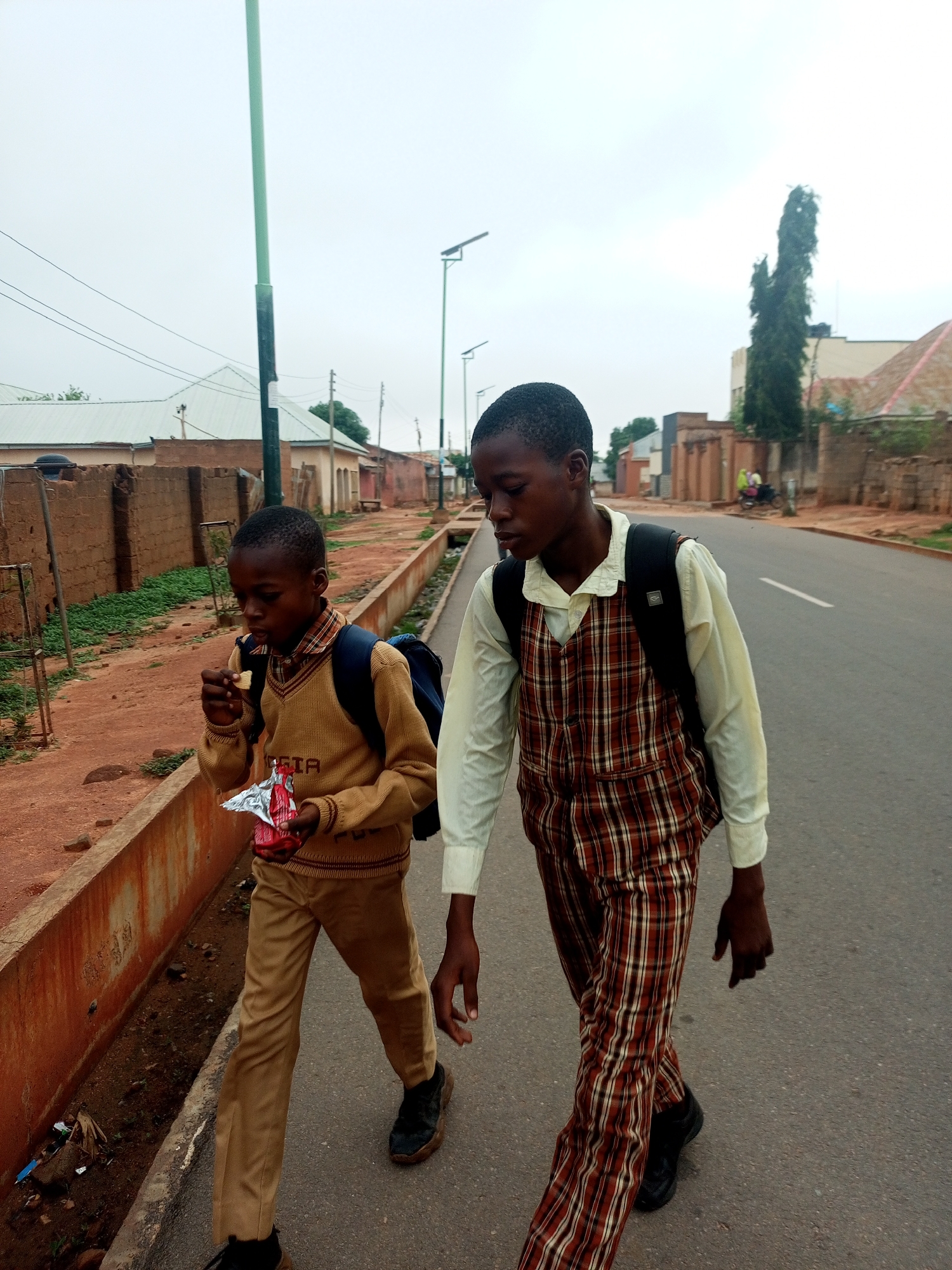 Two young boys, dressed in school uniforms, walk along a paved road in what appears to be a residential area. The boy on the left, wearing a tan sweater vest over a collared shirt and tan pants, is eating a snack from a red and silver wrapper. He has a blue backpack on. The boy on the right, wearing a plaid vest over a cream-colored shirt and plaid pants, also has a black backpack. They are walking on the side of the road, next to a raised concrete curb and a dirt path with some vegetation. In the background, there are buildings made of brick or similar material, and a tall, thin tree. The sky is overcast.