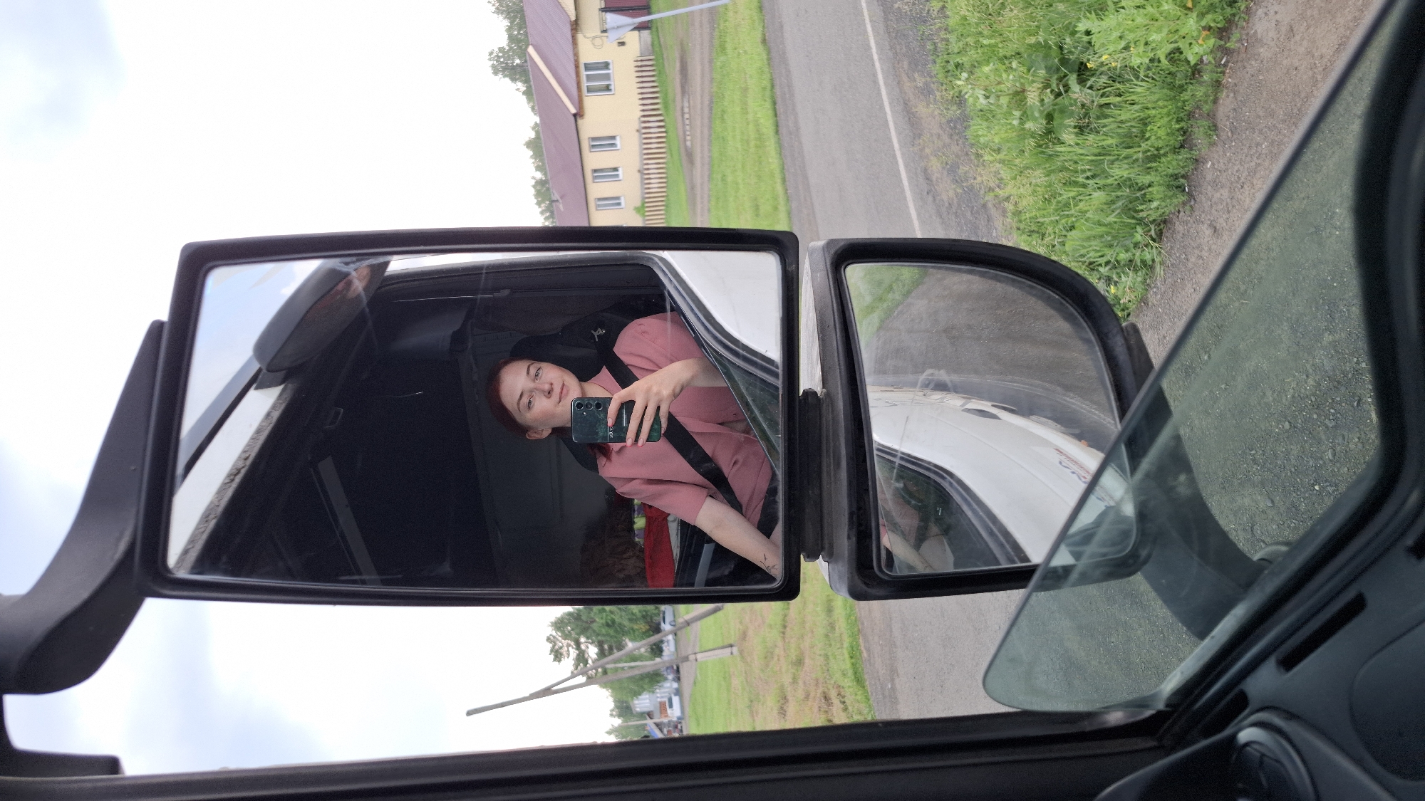 A young woman with short red hair is sitting in the driver's seat of a truck, taking a selfie with her phone. She is wearing a pink collared shirt and has a seatbelt on. The truck's large side mirror reflects her and the interior of the cab. Outside the truck, visible in the mirror, is a rural scene with a road, grassy verge, trees, and a two-story building with a yellow facade and a red roof. The lower mirror reflects the road and the side of the truck as it moves. The scene appears to be in a rural area outside of Nizhny Ingash, Russia.
