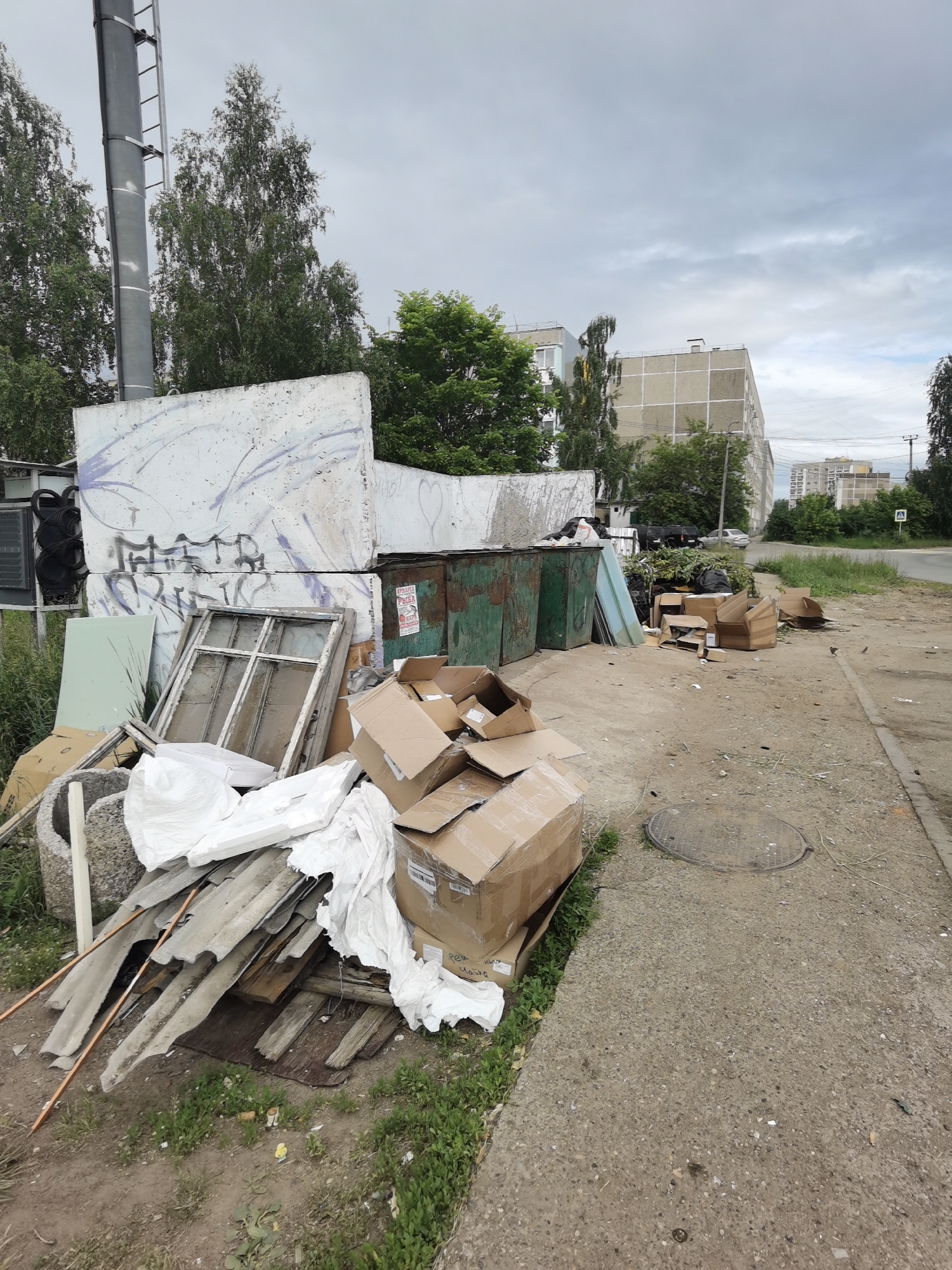 This image depicts a cluttered urban scene in Chaikovsky, Russia, characterized by overflowing dumpsters and discarded materials. In the foreground, a pile of construction debris, including corrugated roofing sheets and wooden planks, is visible, partially covered by white sheeting. Several cardboard boxes, some crushed and open, are stacked nearby. Adjacent to these are two large, green, weathered dumpsters, one of which appears to be filled with refuse. Behind the dumpsters, a large, graffiti-covered concrete structure stands. A broken window frame with glass panes rests against another concrete element. The environment includes a paved sidewalk and a grassy verge, with a manhole cover visible on the pavement. In the background, multi-story residential buildings are present, along with trees and an overcast sky. A lamppost with a ladder is also visible on the left side of the image. The overall impression is one of neglect and disarray.