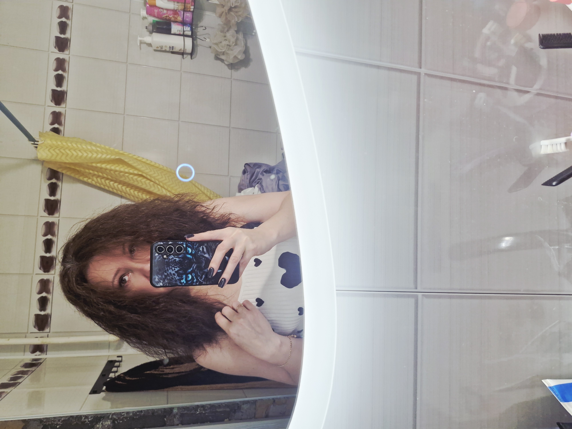 A selfie captured in a bathroom environment, presumably taken in Марковский, Russia. The photographer, a woman with long, brown, wavy hair, is holding a smartphone with a patterned case up to her face. Her reflection is captured in a mirror with an illuminated border. The background reveals bathroom tiles, a shower curtain, and a shelf holding various toiletries. To the right of the mirror's reflection, additional bathroom surfaces and cleaning supplies are visible.