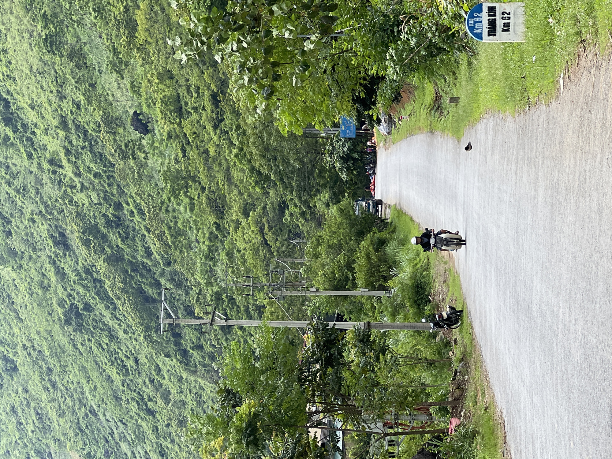 The image depicts a rural road in Cán Tỷ Commune, Vietnam, flanked by lush green, tree-covered mountains. The road is paved and curves gently into the distance. On the left side of the road, there are power lines supported by utility poles, and some residential structures are visible amidst trees. On the right side, more vegetation lines the road, and a white milestone marker indicates "Km 62 TRANG KIM Km 62." A person on a scooter is riding towards the viewer on the right side of the road. Another scooter is parked on the left shoulder. Further down the road, a group of people and several vehicles, including what appears to be a small truck, are gathered. A bird is perched on the edge of the road near the milestone marker. The overall scene suggests a tranquil, natural environment with signs of local life and infrastructure.