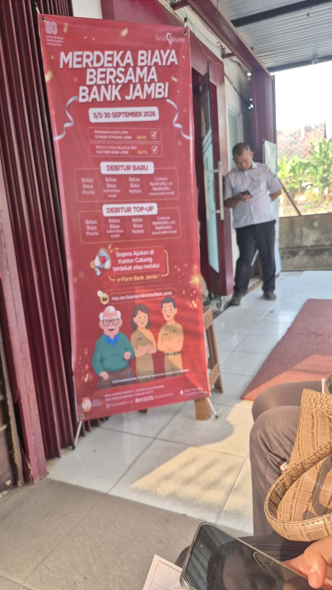 A red banner with white text, advertising a promotion by Bank Jambi, is displayed prominently in front of a building. The banner reads "MERDEKA BIAYA BERSAMA BANK JAMBI" and details various fees that are waived until September 30, 2026, for new and existing debiturers. The promotion includes waived ATM and online transfer fees, as well as cashback offers and waived notary fees for certain loan amounts. The banner also features illustrations of an elderly man and two bank employees, suggesting customer-centricity.
A man in a light-colored collared shirt and dark pants is standing to the right of the banner, looking down at his mobile phone. He appears to be a bank employee, possibly on duty. The environment suggests an outdoor or semi-outdoor setting, with tiled flooring and a building facade in the background. The presence of the banner and the bank employee indicates that this is likely a Bank Jambi branch or promotional event location. The style of the building and the vegetation visible in the background are consistent with a tropical climate, and the mention of "Bank Jambi" strongly suggests the location is in the city of Jambi, Indonesia.