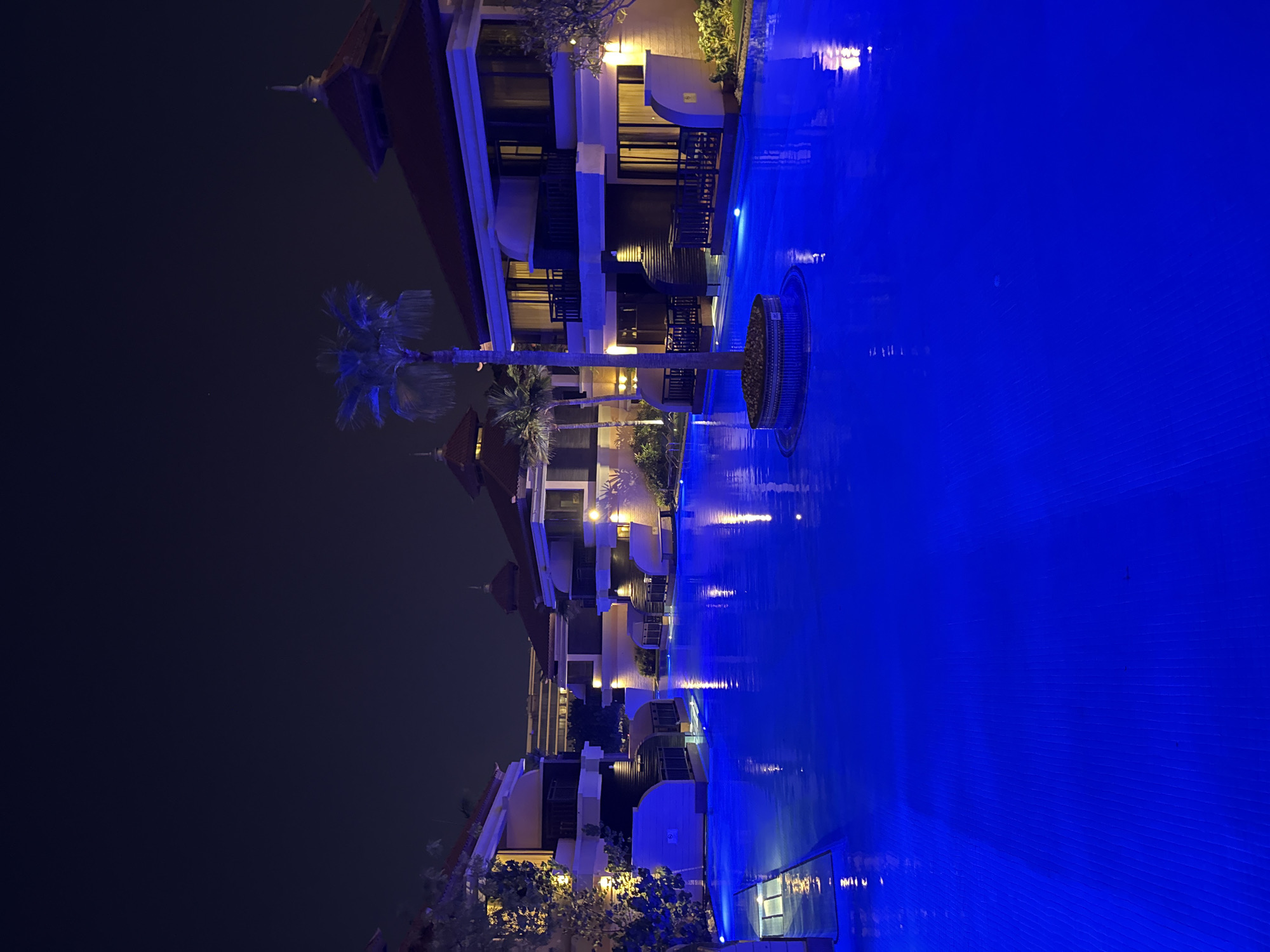 A nighttime exterior shot captures a serene resort swimming pool in Dubai, United Arab Emirates. The water, illuminated by an electric blue light, reflects the surrounding buildings and palm trees. The architecture features traditional elements like pitched roofs and balconies, with warm, yellow lighting emanating from within the rooms. A lone palm tree stands in the center of the pool on a circular island, its fronds silhouetted against the dark night sky. The scene conveys a sense of tranquility and luxury, characteristic of Dubai's upscale hospitality offerings.