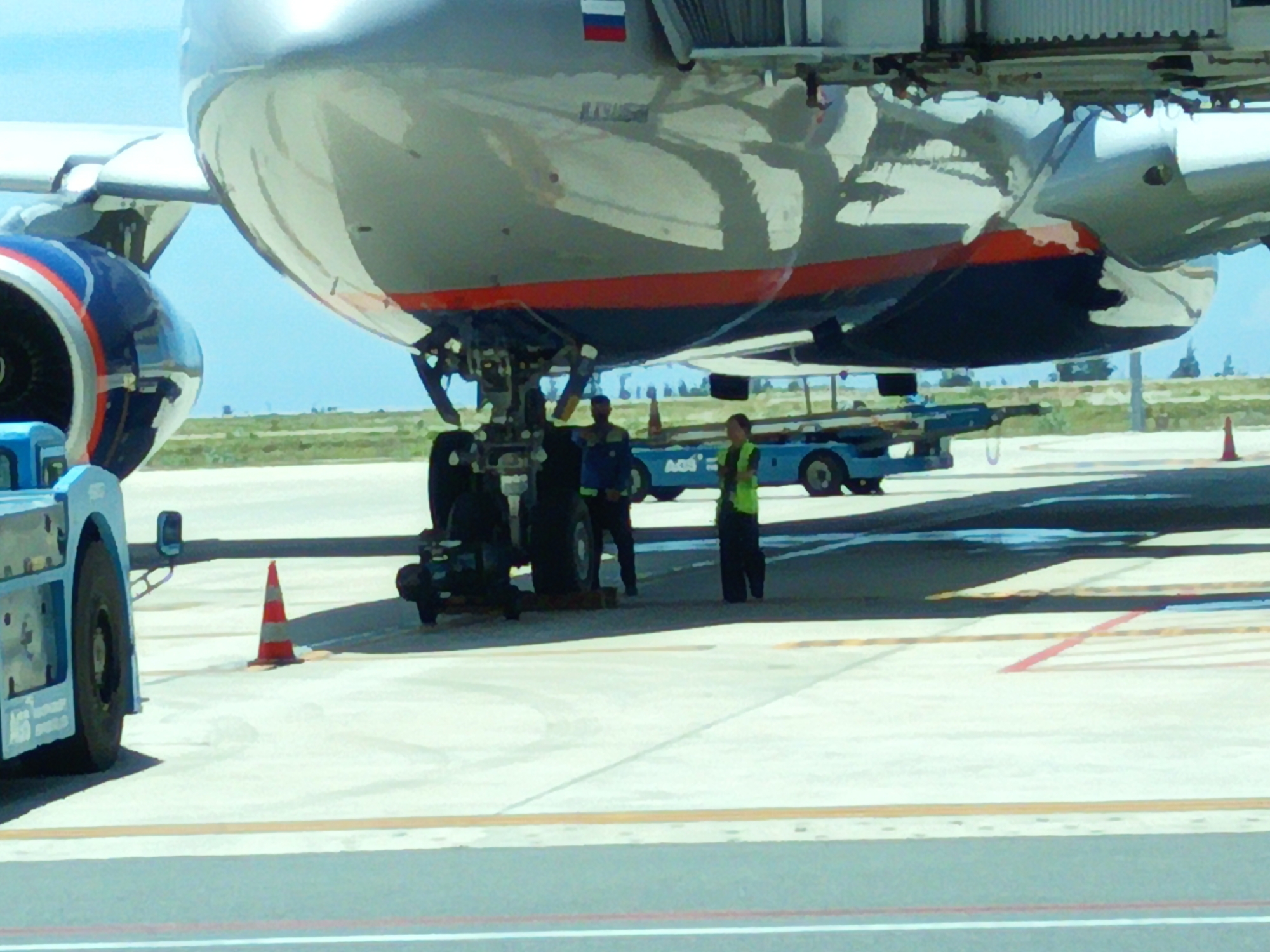 The image shows an airplane on the tarmac at Cam Ranh International Airport in Cam Ranh, Vietnam. The nose of the aircraft is prominent, with a blue and red engine cowling visible on the left. The underside of the plane's fuselage, which is silver with a red stripe, is also visible. Ground equipment, including a blue baggage tractor, is parked near the aircraft. Two individuals, likely airport personnel, are standing near the tractor. One is wearing a blue uniform and the other a high-visibility green vest. A traffic cone is positioned on the tarmac. The sky is a light blue, and the ground is paved with concrete. The overall scene depicts a typical airport environment.