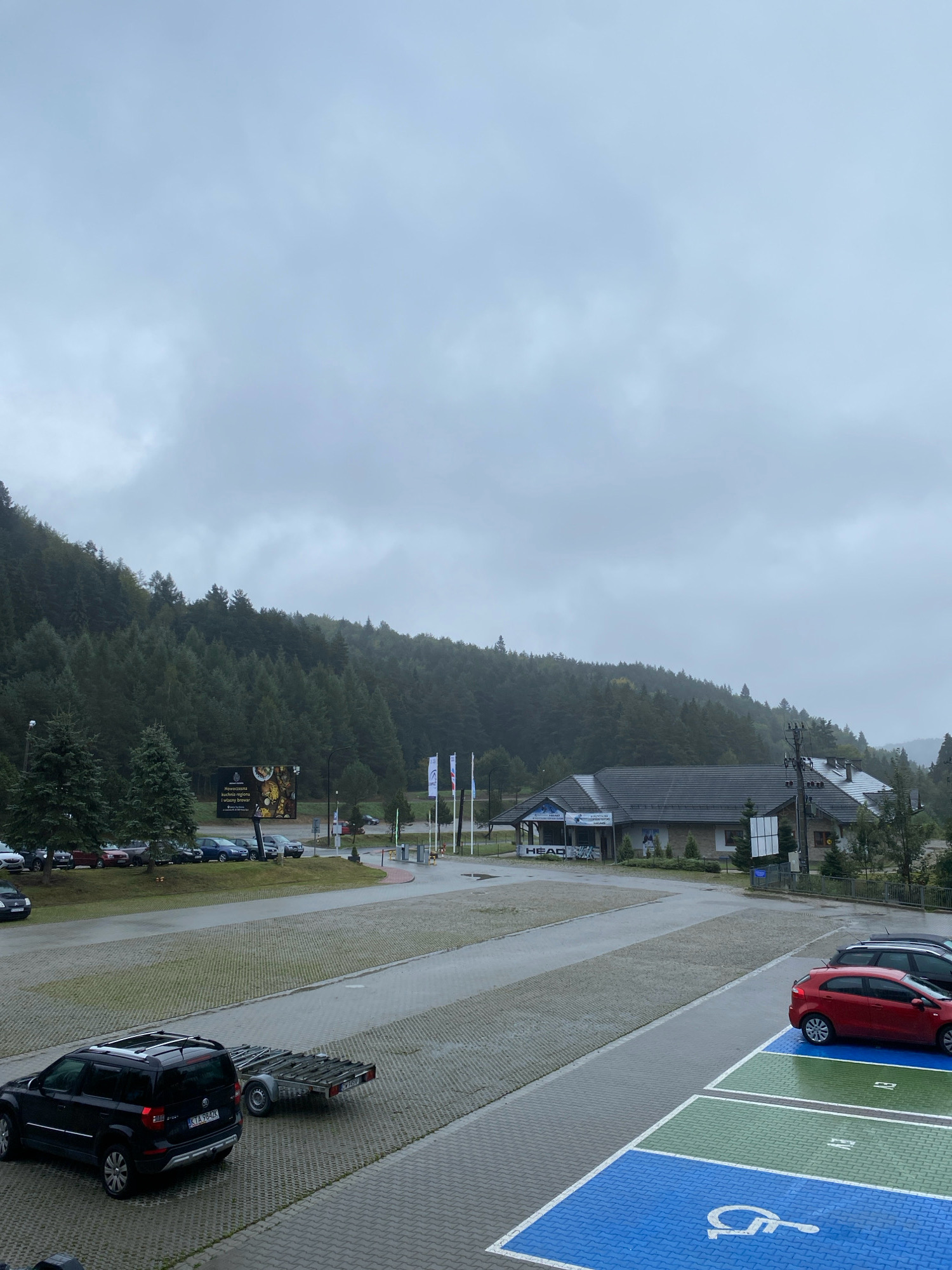 The image depicts an outdoor parking lot in Krynica-Zdrój, Poland, under a cloudy sky. In the foreground, a black SUV with a trailer attached is parked on a gray paved surface. To the right, a red car is parked in a designated handicapped spot marked with blue and green paint and a wheelchair symbol. Beyond the parking lot, there is a road and a building with "HEAD" signage. Further in the background, a dense forest covers a hillside. A large billboard is visible among the trees, displaying an advertisement in Polish. The overall scene suggests a somewhat overcast day in a natural, mountainous area, likely a tourist destination. There are no people visible in the image.
