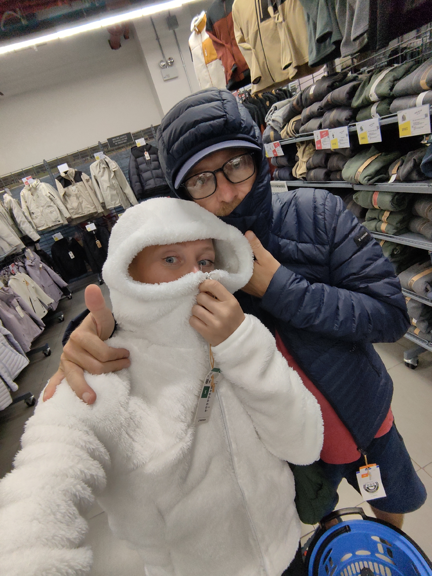 This photograph captures a lighthearted moment between two individuals, likely an adult and a child, inside a clothing store in Hà Nội, Vietnam. The scene suggests a shopping trip for outerwear, possibly at a Decathlon store given the brands visible.
In the foreground, a child, whose gender is not explicitly clear but appears to be female, is wearing a fluffy white hooded jacket with the hood pulled up high, obscuring most of their face except for their bright blue eyes and nose. Their left hand holds the jacket up around their face in a playful gesture, while their right hand gives a thumbs-up. To the right of the child, an adult male with glasses and a beard is partially visible, wearing a dark blue hooded puffer jacket with a "S SIMOND" logo on the sleeve. He looks towards the camera with a gentle expression, indicating a selfie.
The setting is a well-lit retail space, characterized by multiple racks and shelves stocked with various jackets and coats in the background. The clothing items are neatly displayed, some hanging and others folded. The style of jackets suggests an outdoor or sports apparel store. A blue shopping basket handle is partially visible in the bottom right corner, reinforcing the shopping context.
Notable details include the playful and joyful mood conveyed by the subjects. Visible text on a sign in the background reads, "MỜI THỬ JACKETS NÀY VÀ CÁC LOẠI ĐỘ ĐỘC ĐÁO VÀ THỂ THAO ĐI BỘ," which translates from Vietnamese to "Try these jackets and unique items for sports and hiking." Price tags on the jackets show figures like "399k," "419k," and "299k," likely indicating prices in thousands of Vietnamese Dong. Another tag near the basket shows "B'TWIN," a Decathlon brand for cycling. The overhead fluorescent lighting suggests an indoor commercial environment.