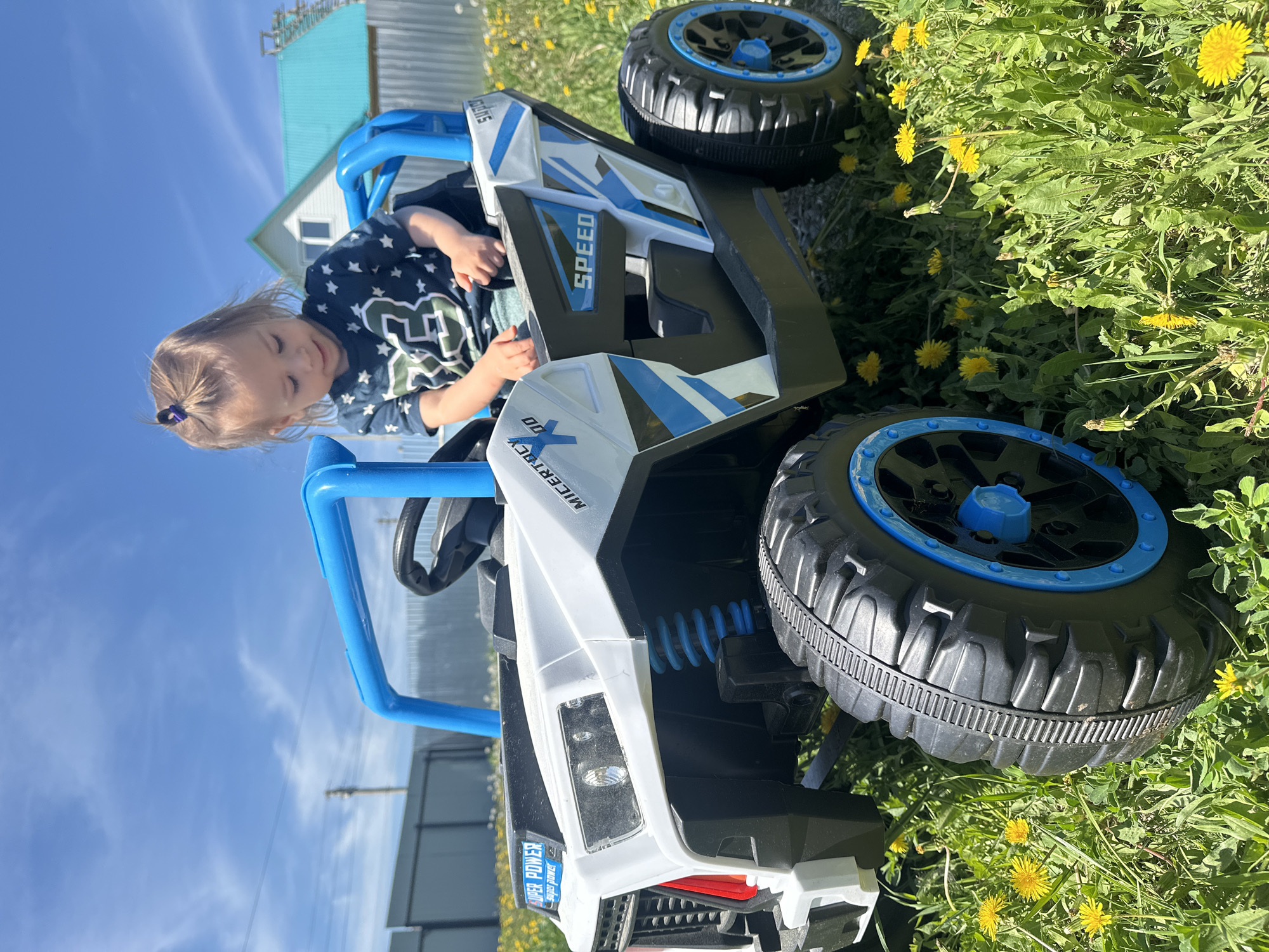 A young child is seated in a toy off-road vehicle on a grassy lawn with yellow dandelions. The vehicle is predominantly white, black, and blue, with "SPEED" written on its side. A building with a light blue roof is visible in the background, along with a fence and clear blue sky. The child is wearing a dark blue shirt with white stars and a green number. The location is Kharnavy, Russia.