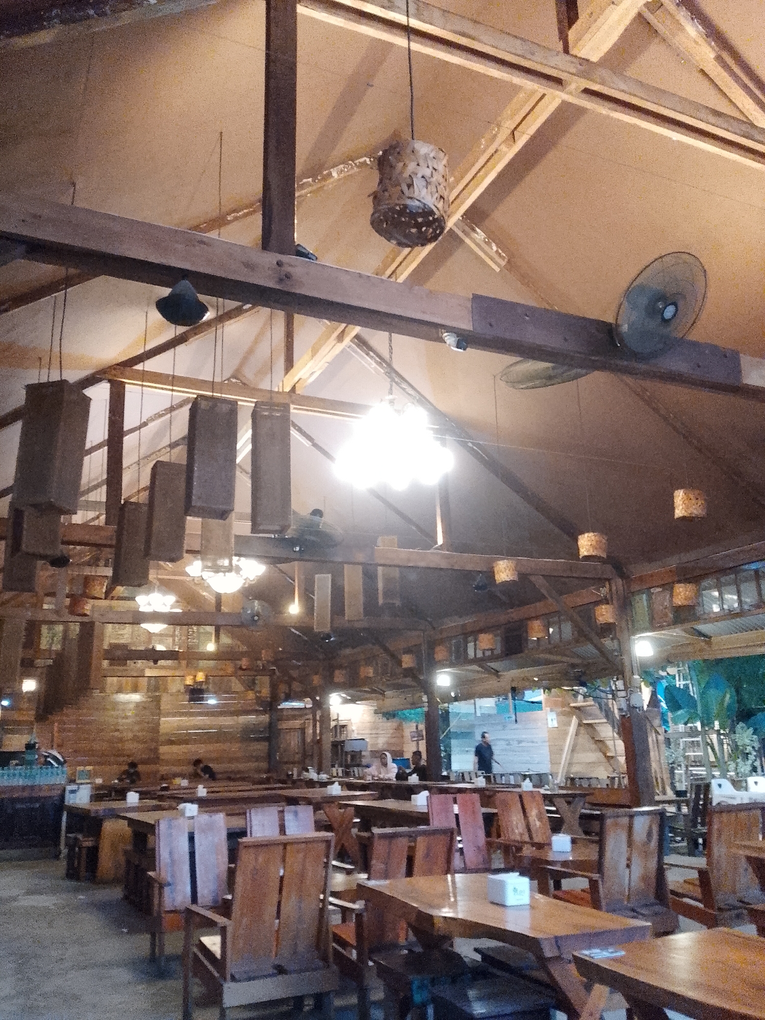 The image depicts the interior of a restaurant, presumably in Lhokseumawe, Indonesia. The dining area is furnished with wooden tables and chairs, some occupied by people. The roof structure is exposed timber, with hanging lights and fans. The walls are made of wood. Some people can be seen in the background. Outside, some green plants are visible.