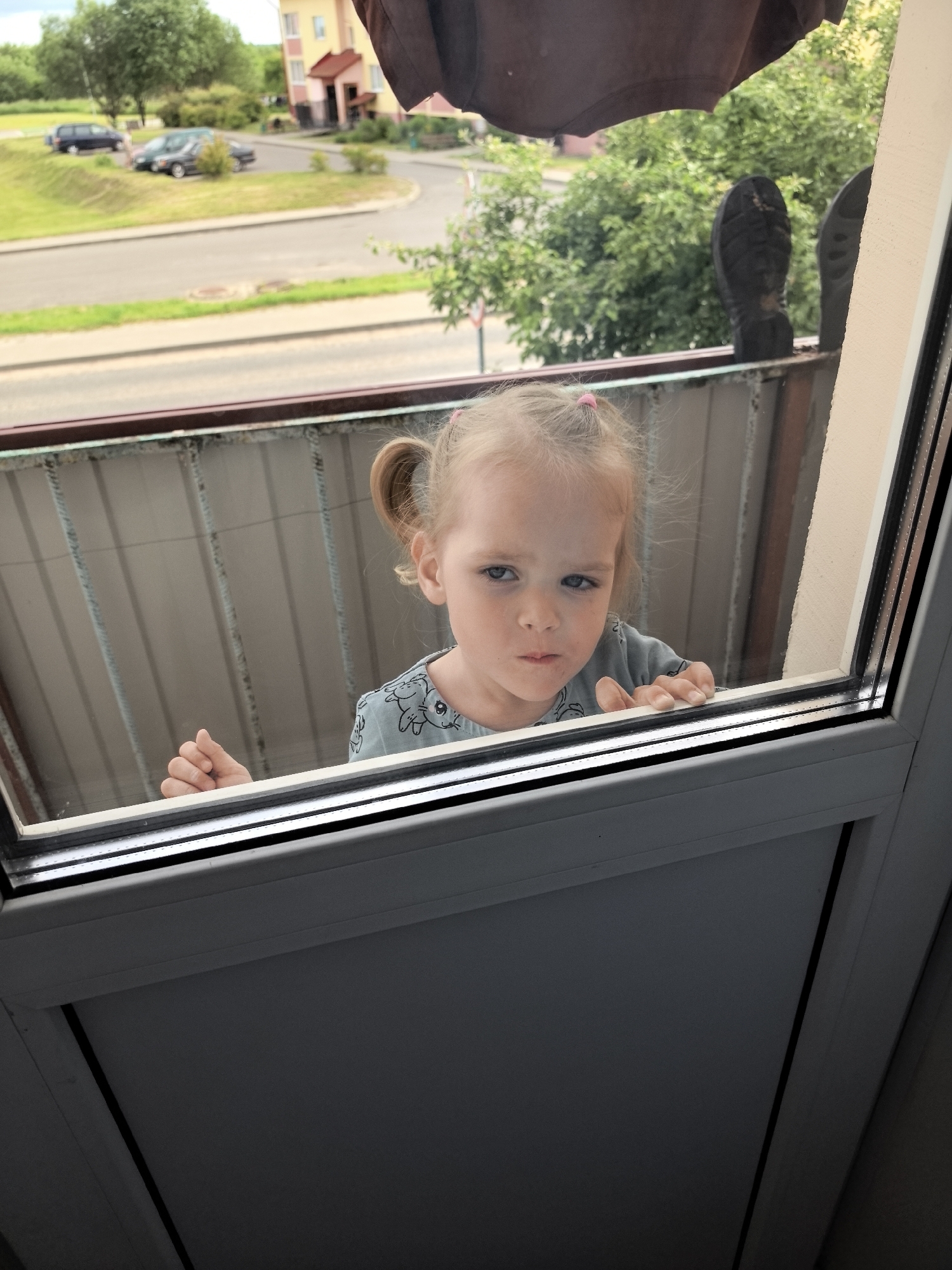 A young child with blonde hair in two pigtails is looking through a window. The child is wearing a light blue t-shirt with a repeating pattern of cartoonish animals. Their face shows a slightly unhappy or grumpy expression. Their left hand is resting on the window frame, with fingers visible.
The scene appears to be from a balcony, looking out onto a street with some cars and trees. In the background, there is a building with a yellow facade and a red roof. The weather appears to be overcast, as indicated by the diffused lighting. The time of day is likely daytime. The child is positioned on a balcony with a metal railing and a corrugated metal wall behind them. There are also two black shoes resting on the railing, with their soles facing the camera, suggesting someone is standing on the balcony or has placed them there. The overall setting seems to be a residential area. There is no visible text in the image.