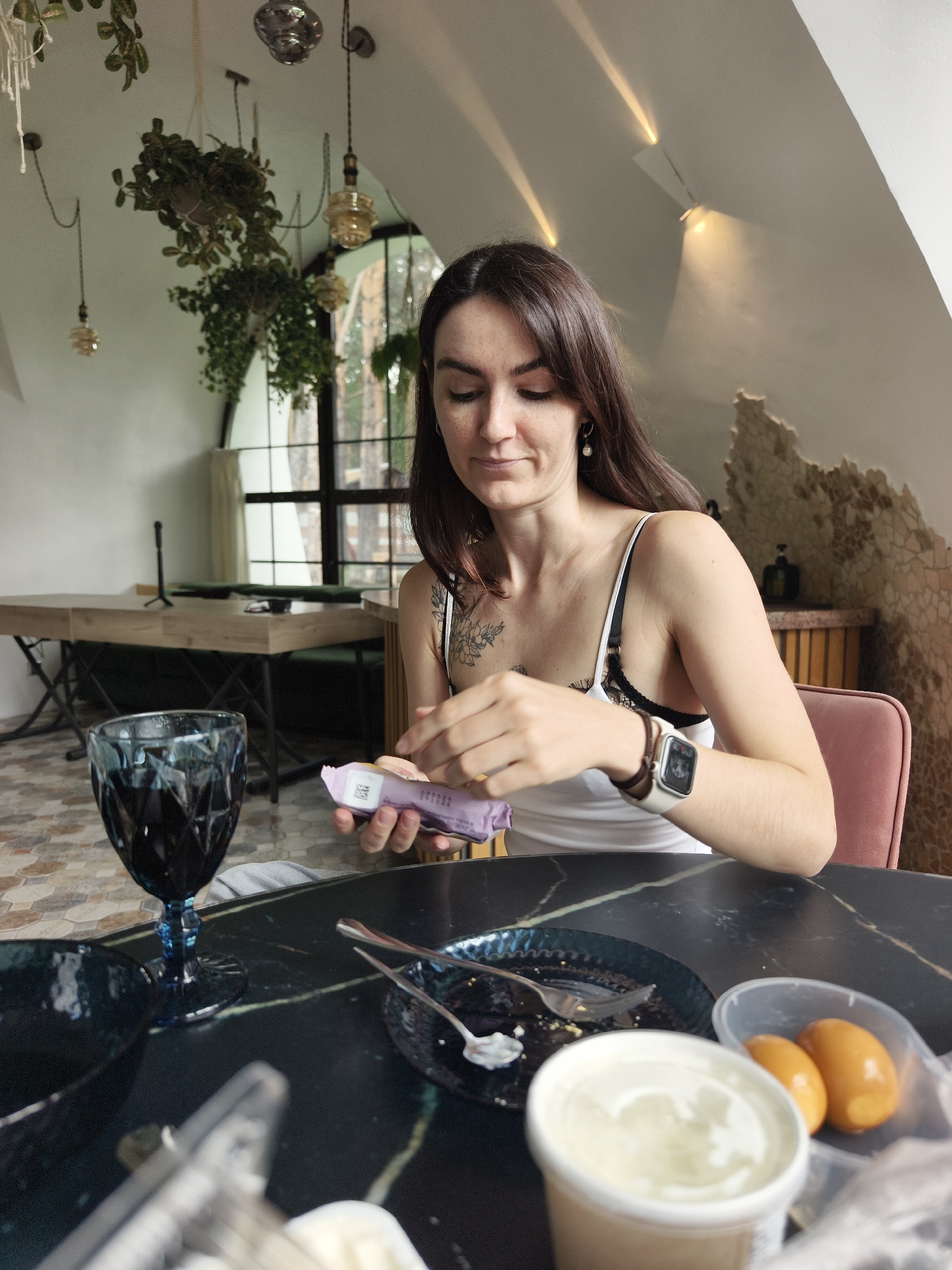 A woman with long dark hair is seated at a dark marble table in what appears to be a cafe or restaurant. She is wearing a white tank top with black lace trim and a smartwatch on her left wrist. She is holding a purple wrapper in her hands, appearing to open it. In front of her is a dark blue faceted glass filled with a dark liquid, a small blue plate with remnants of food and cutlery, a white container of what looks like yogurt or sour cream, and a plastic bag containing two boiled eggs. The background reveals a large window with a dark frame, offering a glimpse of greenery outside. Hanging plants and decorative light fixtures are visible on the ceiling. The overall ambiance suggests a relaxed dining setting. The location is identified as OnoKhino, Russia.