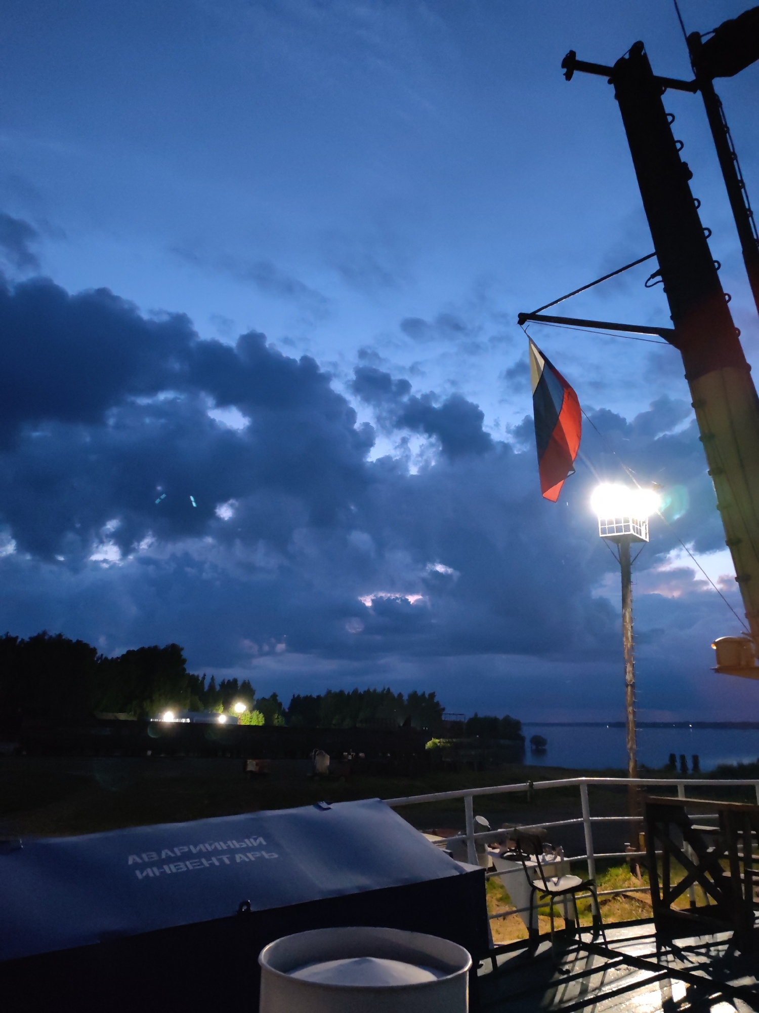 The image depicts a view from a vessel in the city of Rybinsk, Russia, during twilight. A Russian flag flies from a mast in the foreground, illuminated by a nearby lamp. In the background, a body of water stretches towards a tree-lined shore with scattered lights. A railing is visible in the mid-ground, along with some outdoor furniture and a covered object with Russian text that reads "аварийный инвентарь" (emergency inventory). The sky is a deep blue with dramatic, dark clouds.