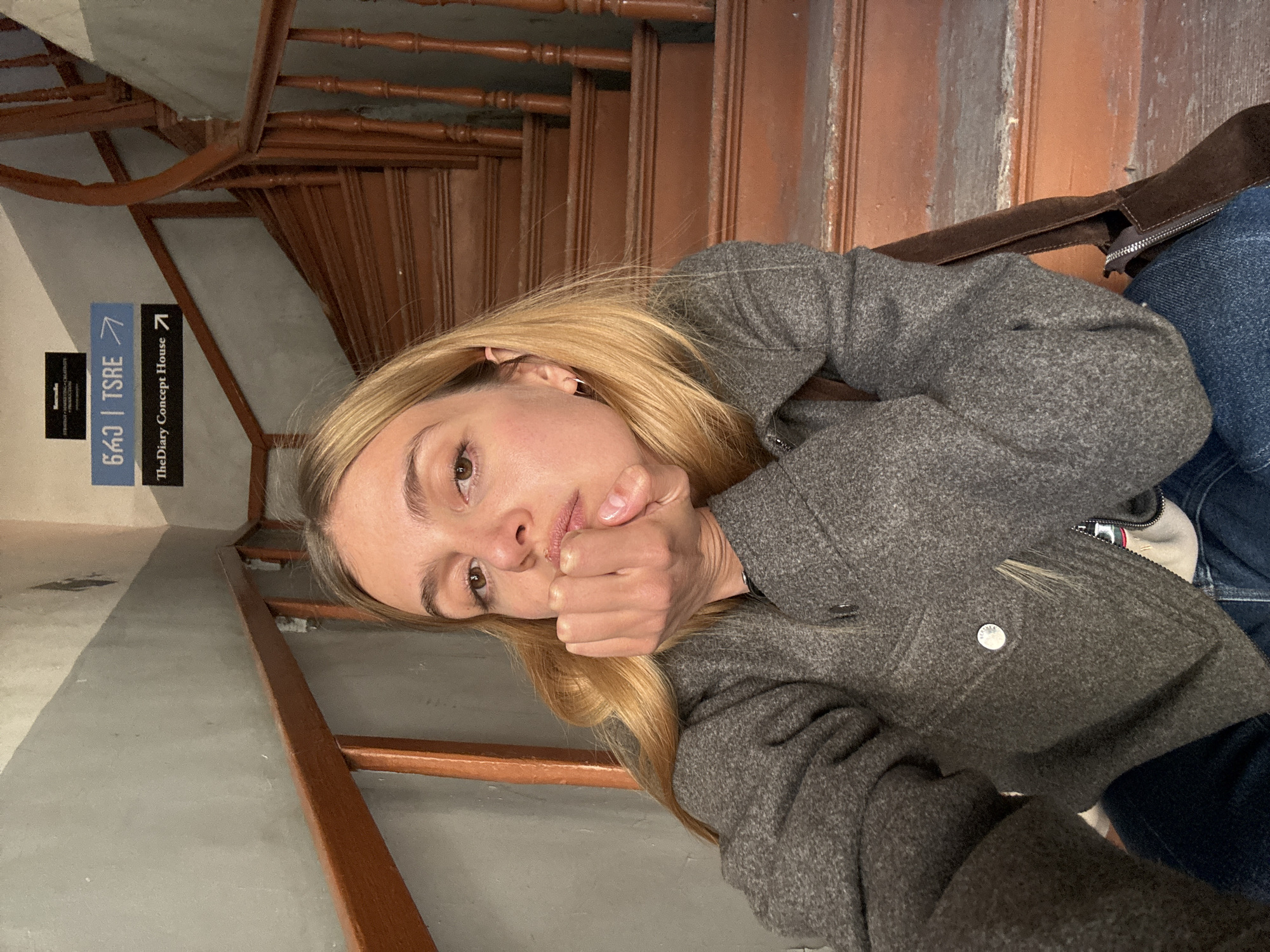 The image shows a blonde woman sitting on a wooden staircase, possibly in Tbilisi, Georgia, based on the presence of a sign that says "TheDiary Concept House" and "643 Tsre". The woman wears a grey coat and blue jeans and rests her chin on her hand. A brown suede shoulder bag is partially visible to her right. The background features the staircase, and a wall with a sign.