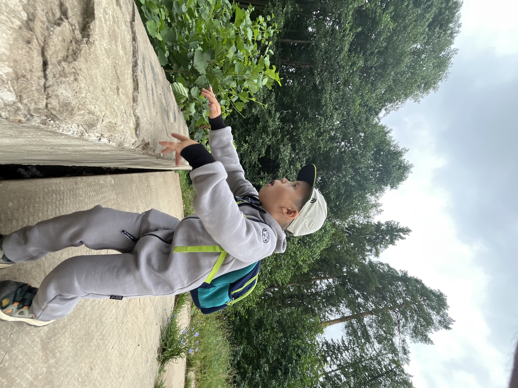 In Chaikovsky, Russia, a young boy wearing a grey tracksuit, a white baseball cap, and a blue and green backpack leans over a concrete barrier. He is reaching out with his hands as if to touch something. Lush green foliage is visible on the left, and trees form a dense backdrop against a cloudy sky. The ground is a concrete pathway.
