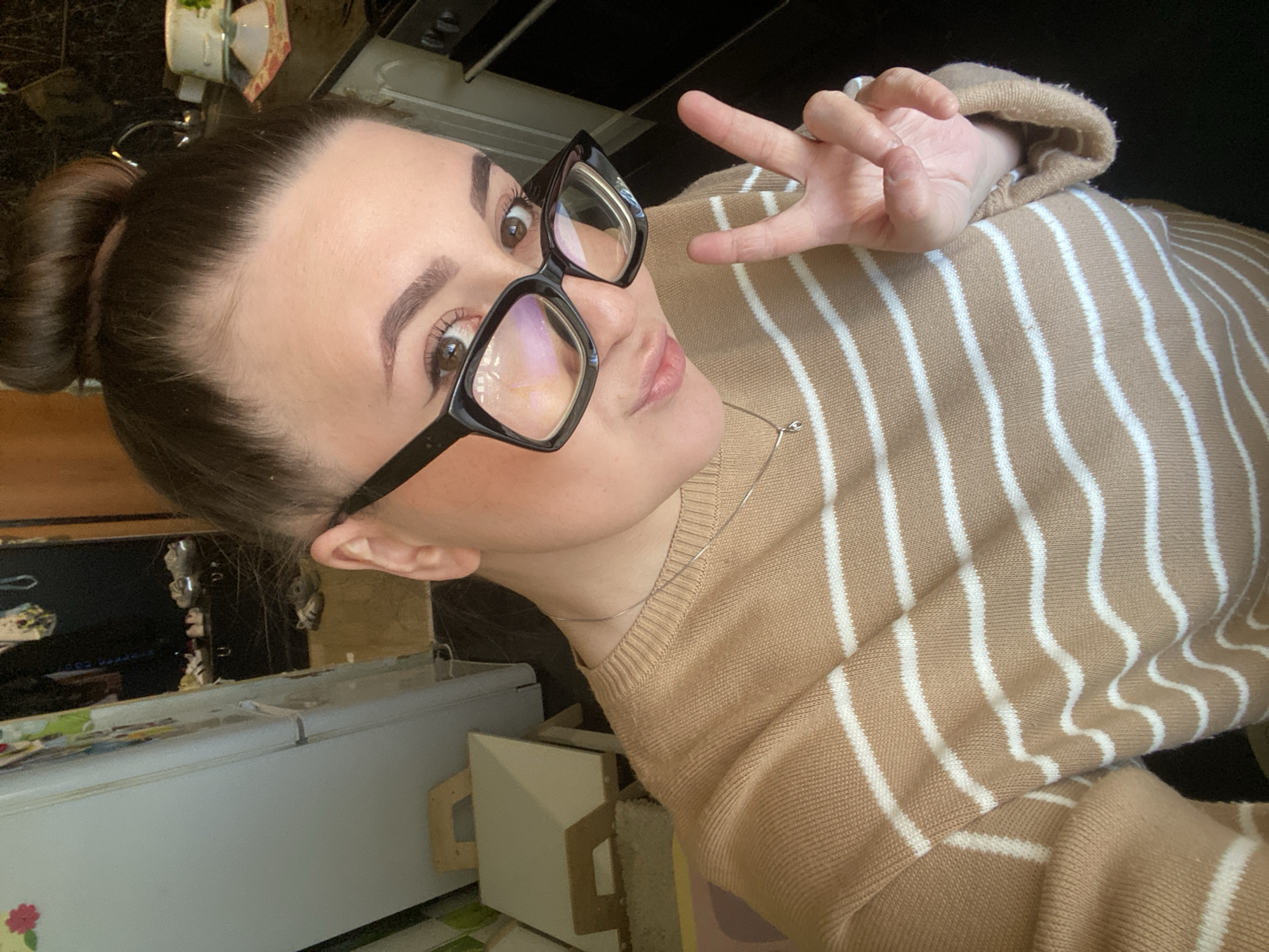 A young woman with brown hair in a bun is taking a selfie indoors. She is wearing black-framed glasses and a tan and white striped sweater, holding up a peace sign with her right hand. The background includes kitchen elements like a countertop, cabinets, and a refrigerator. The location cannot be determined to be Chaikovsky, Russia from the image.
