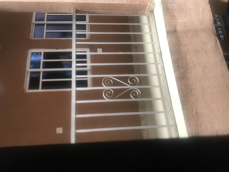 The image shows a close-up view of a window with a white metal grate in front of it. The grate is made of horizontal bars and features a decorative design at the bottom. The window is divided into two sections, with the top section having a small space between each pane of glass and the bottom section having a larger space between each pane. The window is on a brown wall, with a light brown trim. The building is located in Umudike, Nigeria, at the coordinates 5.471° N, 7.543° E. There are no people or objects visible in the image, apart from the window and grate. The environment is typical of an urban setting. The scene is stationary, and there is no action taking place.