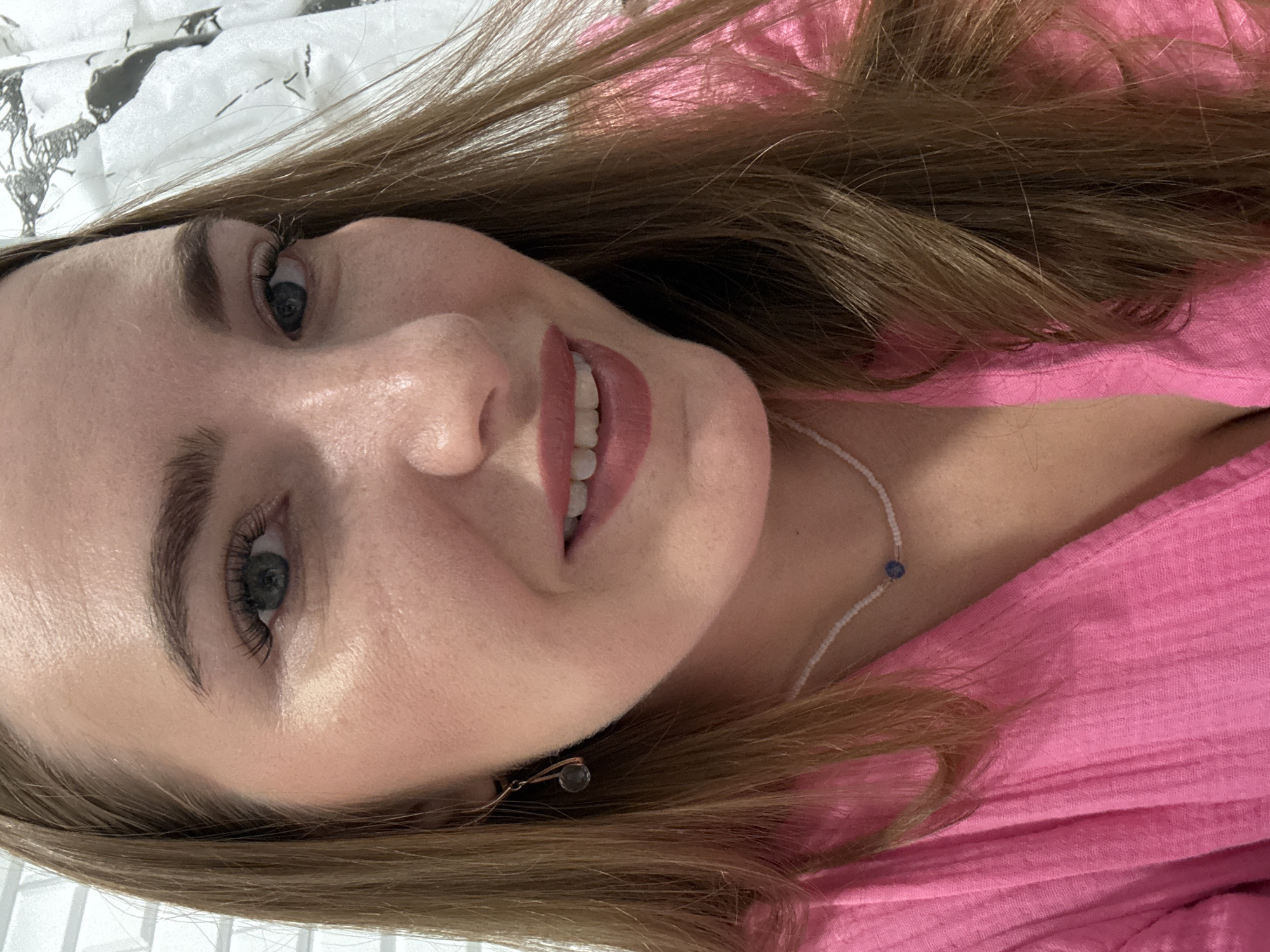 A close-up, head and shoulders, natural light portrait of a young Caucasian woman with light brown hair, green eyes, and fair skin. She is wearing a bright pink, textured V-neck top and a delicate white beaded necklace with a single blue pendant. A small, dark teardrop-shaped earring is visible on her left earlobe. She is smiling with her teeth showing. The background is out of focus and appears to be a light-colored, textured surface, possibly a curtain or wall covering with a patterned design. The lighting suggests an indoor setting.