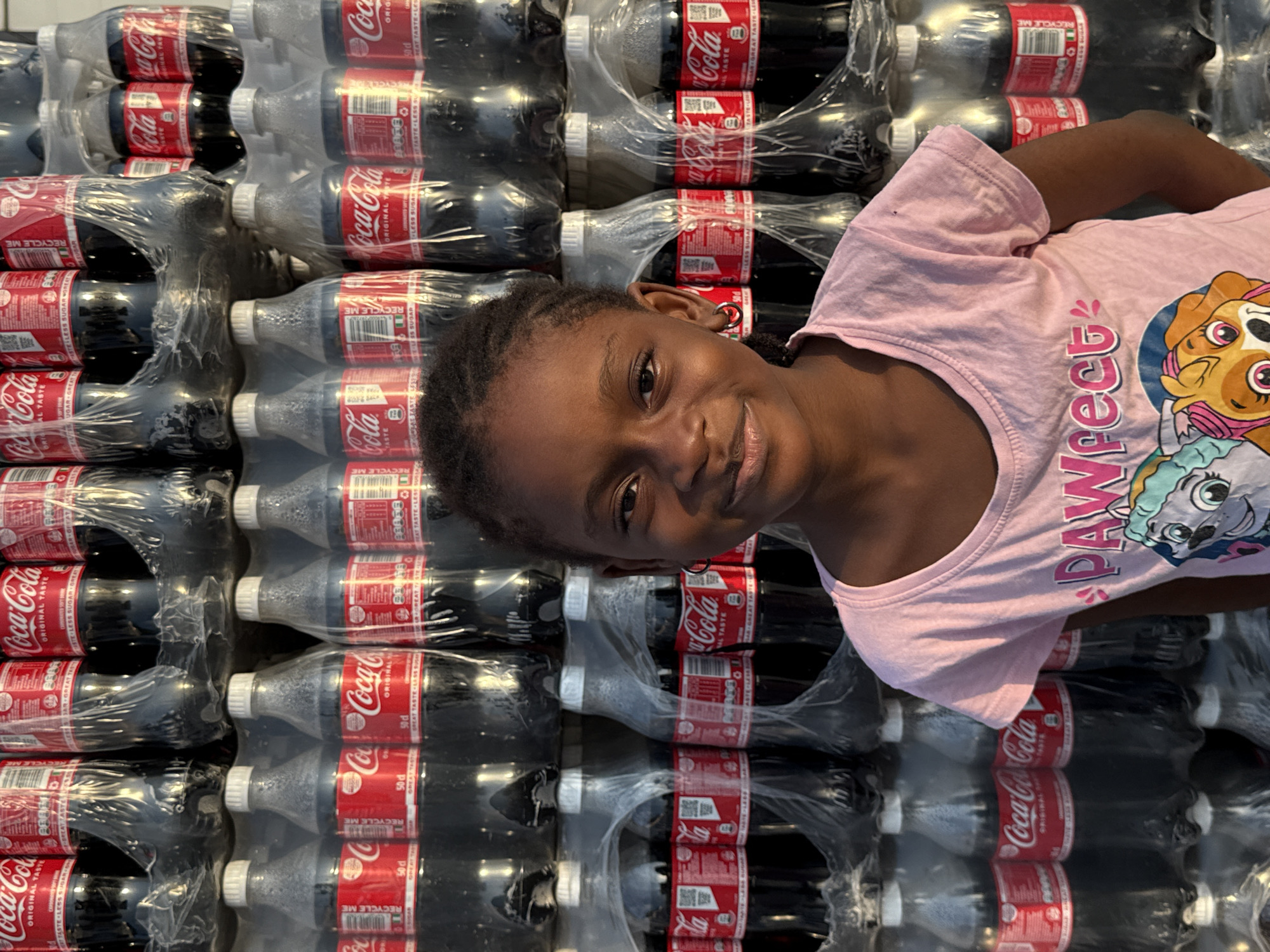A young Black girl with braided hair and wearing a pink t-shirt with cartoon characters on it is standing in front of a large stack of Coca-Cola bottles. The bottles are arranged in multiple rows and are wrapped in plastic. The background suggests an outdoor setting in a city, possibly Ado, Nigeria, given the context of the provided information. The girl is looking directly at the camera and smiling slightly. The lighting indicates it is daytime, with some shadows suggesting the sun is overhead or to the side. The image is taken from a slightly low angle, emphasizing the height of the Coca-Cola stack.