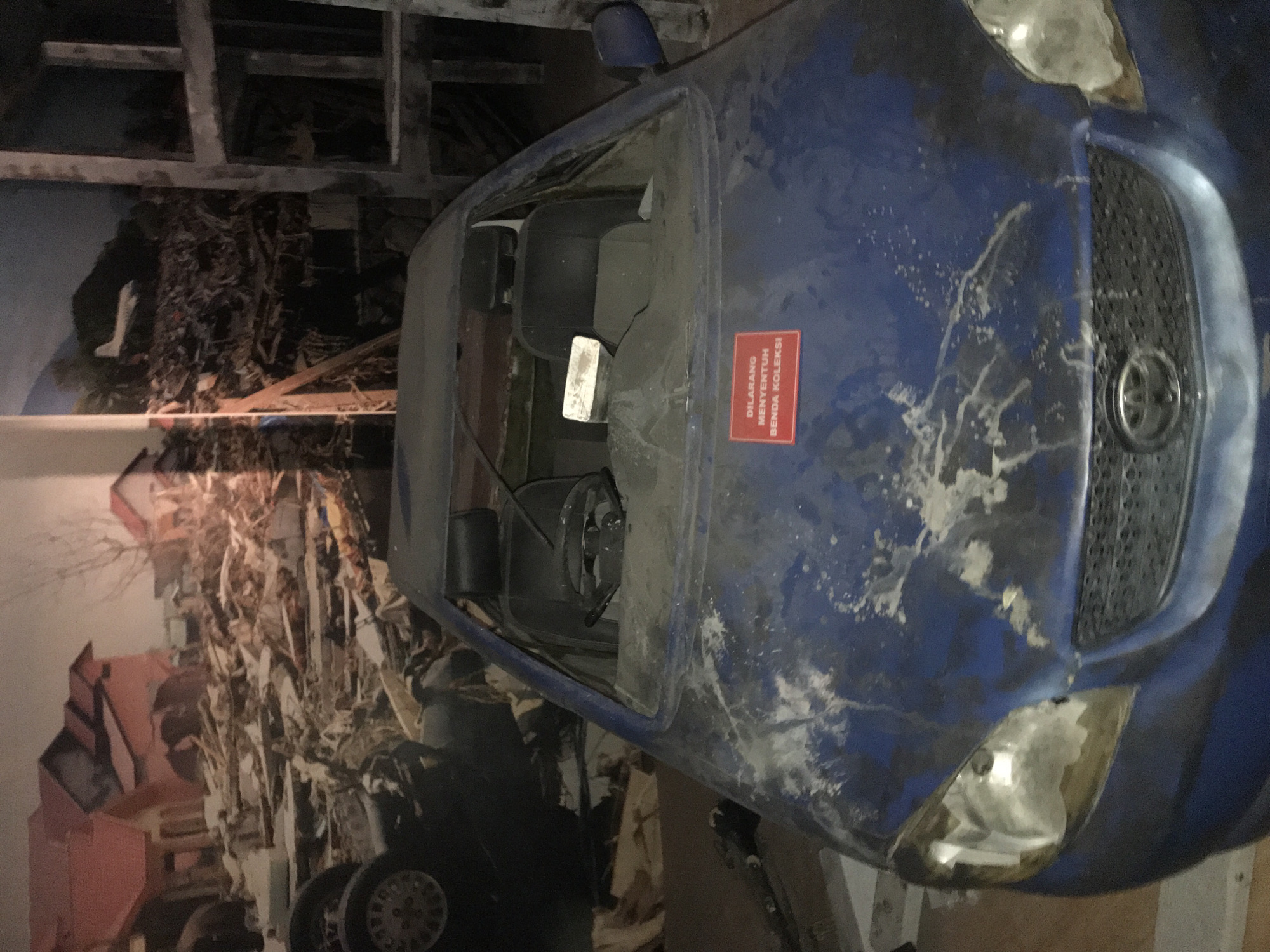 The image depicts a blue Toyota Corolla, heavily damaged and covered in debris, on display against a backdrop of destruction. The car's windshield is missing, and its body is marred with scratches and dried mud or silt. A red sign in Indonesian is affixed to the dashboard. The car's front end, including the grill and headlights, is visible. The background shows a mural or large photograph of the aftermath of a disaster, likely a tsunami, depicting destroyed buildings and debris. Based on the car's display and the imagery in the backdrop, the image likely represents a memorial or exhibit related to the 2004 tsunami in Banda Aceh, Indonesia. The environment is a museum.