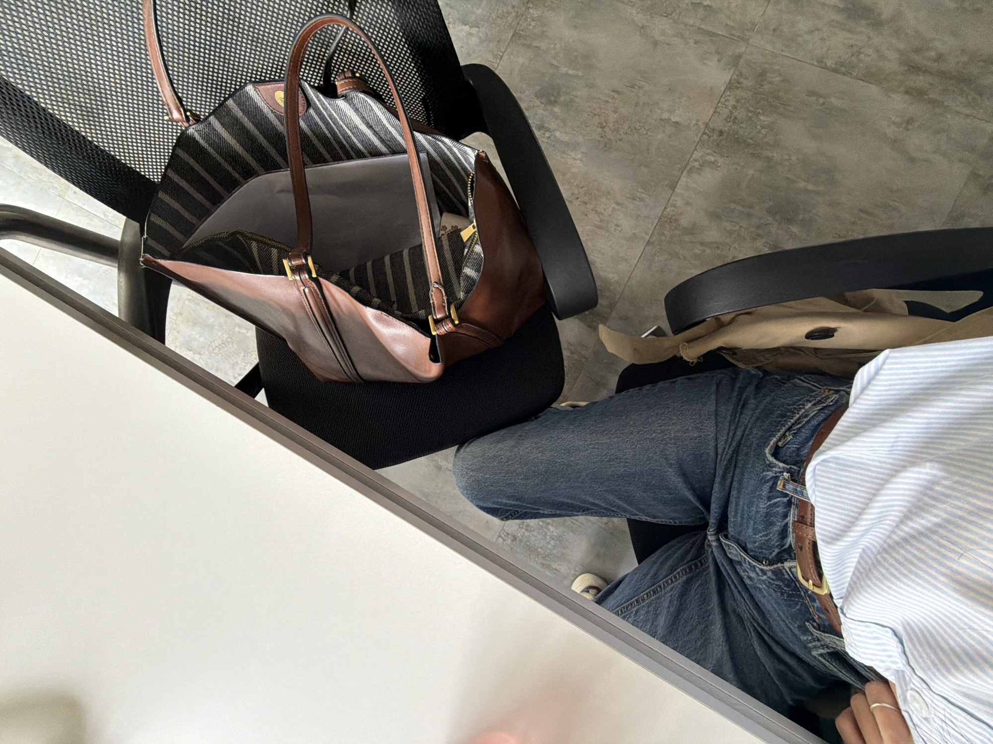 The image shows an office setting. A black mesh office chair holds a brown leather handbag with striped lining. Beside this chair is another black chair with a beige jacket. A person wearing blue jeans, a brown belt, and a striped shirt is sitting in the chair. The scene includes a white desk and tiled flooring. The location in Yerevan, Armenia cannot be determined from the image.