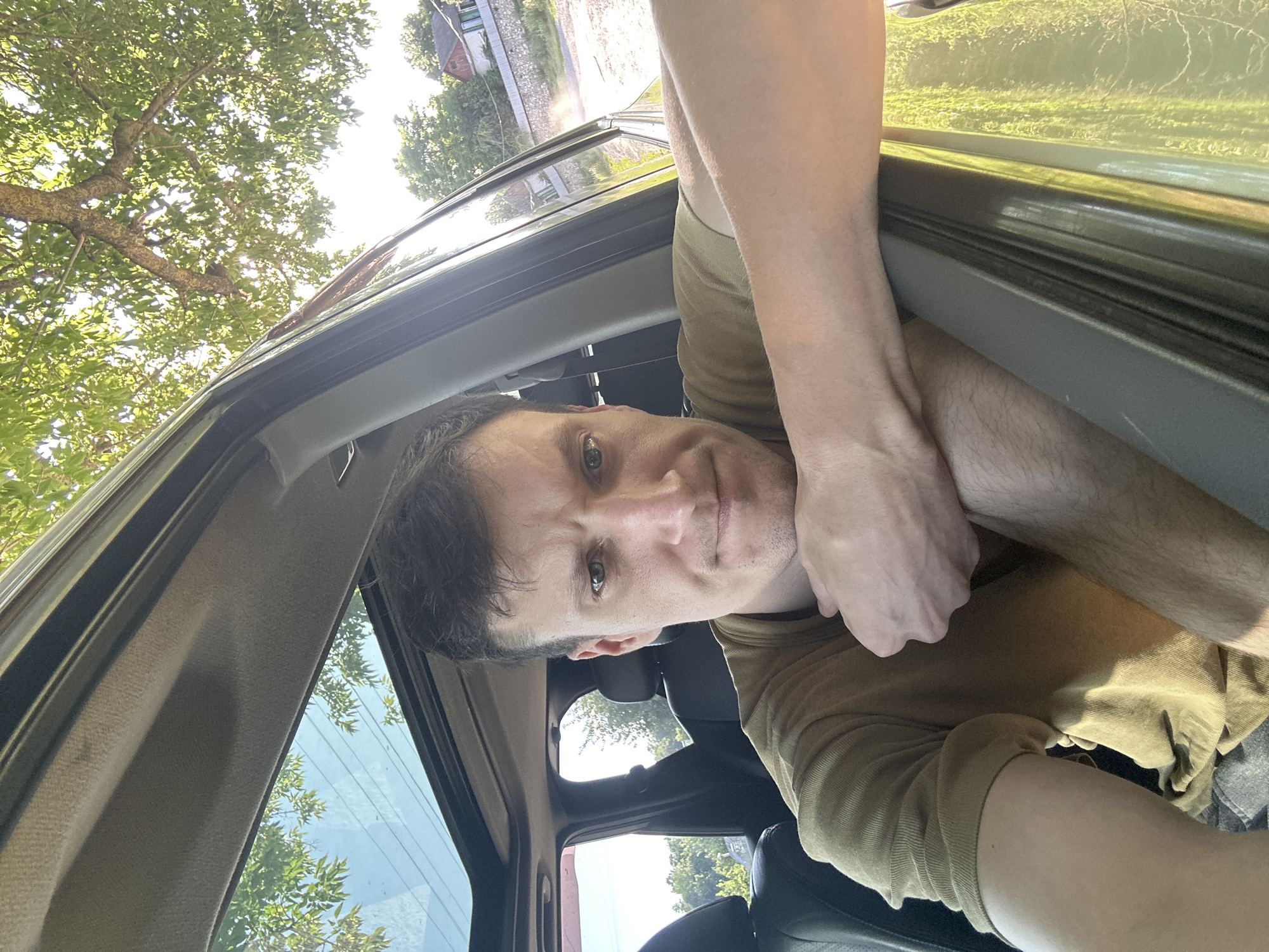 The image is a selfie taken from inside a car. A man with short brown hair and green eyes is leaning his arm on the open car door. He is wearing a tan t-shirt. The car has a sunroof with the shade pulled back, revealing a clear blue sky and green tree branches. The background shows trees, buildings, and a stone wall, suggesting an outdoor setting in a semi-rural or suburban area. The lighting indicates it is daytime. While the image was provided with context referencing the city of Makiv, Ukraine, the visual elements within the photograph itself do not contain specific landmarks or identifiers that definitively confirm this location.