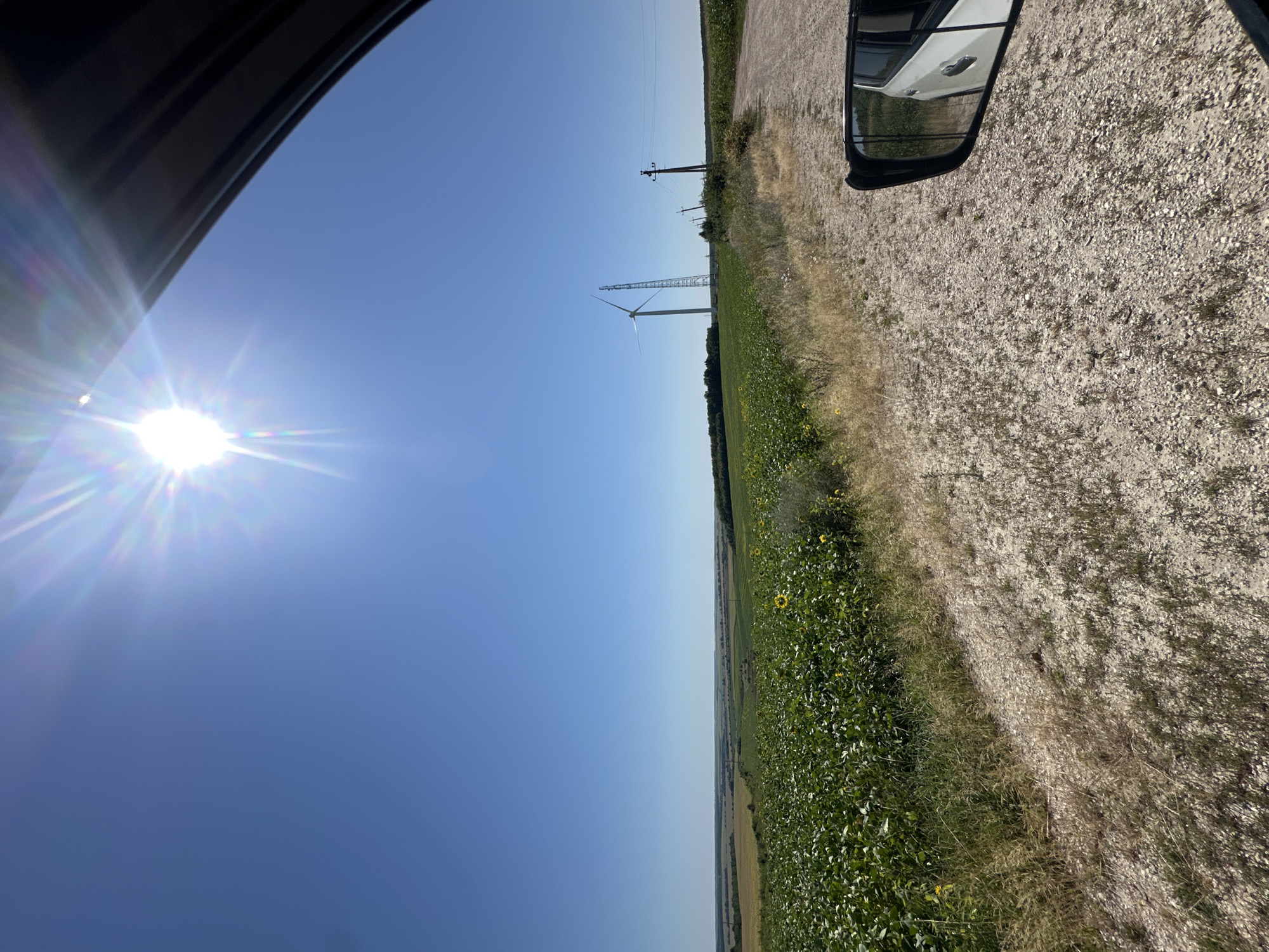An eye-level, wide shot captures a sunny day in Korchivka, Ukraine, with a view from the passenger side of a vehicle. The sky is a clear, bright blue, dominated by the sun's intense glare in the upper left quadrant, casting starburst rays. In the middle ground, a vast field of lush green crops, possibly sunflowers, stretches across the landscape. Beyond the crops, a single white wind turbine stands tall, accompanied by a lattice tower structure. To the right, a gravel road curves into the distance, bordered by power lines and poles. The side mirror of the car reflects a white vehicle and the surrounding greenery. The foreground consists of a dirt and gravel road with sparse dry grass.