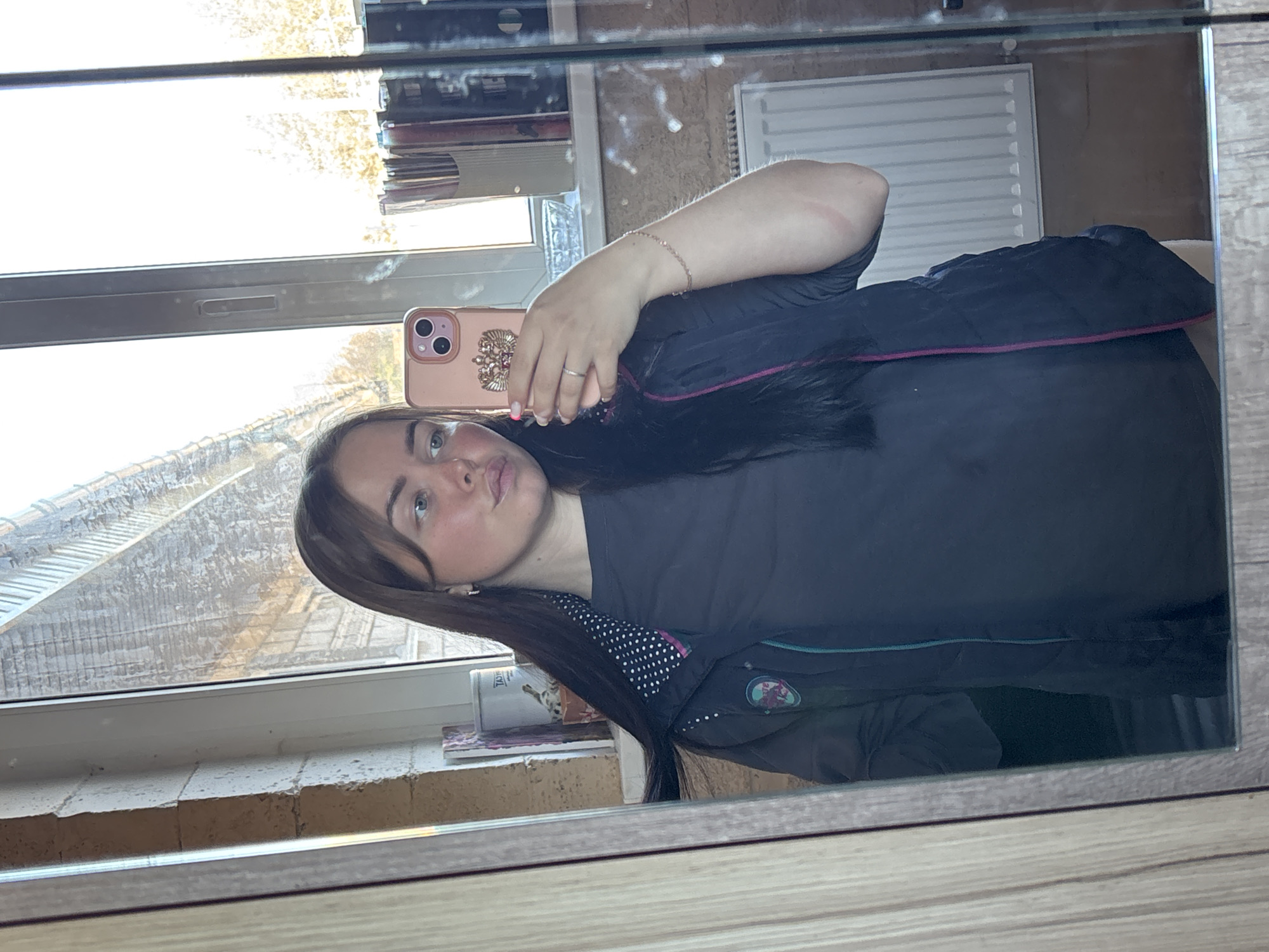 A young woman takes a selfie in a mirror. She holds a rose-colored phone case. She is wearing a black t-shirt and a black puffy jacket. The mirror reflects an interior space, including a window with a view of buildings and trees, and a radiator. The image gives no clues about the location being Русский Вожой, Russia.