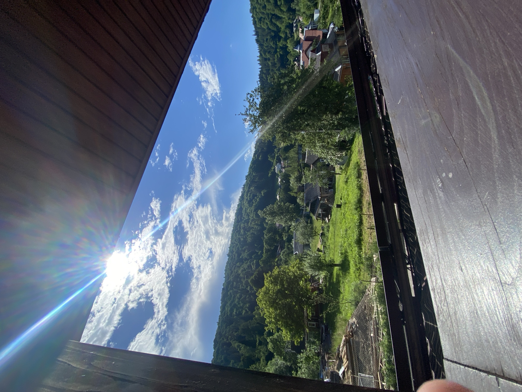 This is a vertical, medium-distance, eye-level shot taken outdoors, looking out from under a dark, sloped roof structure. The view encompasses a sun-drenched, verdant rural landscape in Yaremche, Ukraine. The sun is prominently positioned in the upper left quadrant, casting brilliant rays and a lens flare across the bright blue sky dotted with fluffy white clouds. Below the sky, a lush green hillside is densely covered with various shades of green trees, sloping downwards. Scattered amongst the trees are several houses with red tile roofs, characteristic of the region. In the foreground of the landscape, closer to the viewer, are more houses, some appearing to be under construction with visible wooden planks and piles of materials. A bright green, grassy field with a few scattered trees dominates the middle ground. The roof structure from which the photo is taken is dark brown and textured, with visible wooden beams and a darker, sloped surface in the foreground. The overall scene depicts a bright, sunny day in a peaceful, hilly, and wooded area with signs of both established and ongoing development.