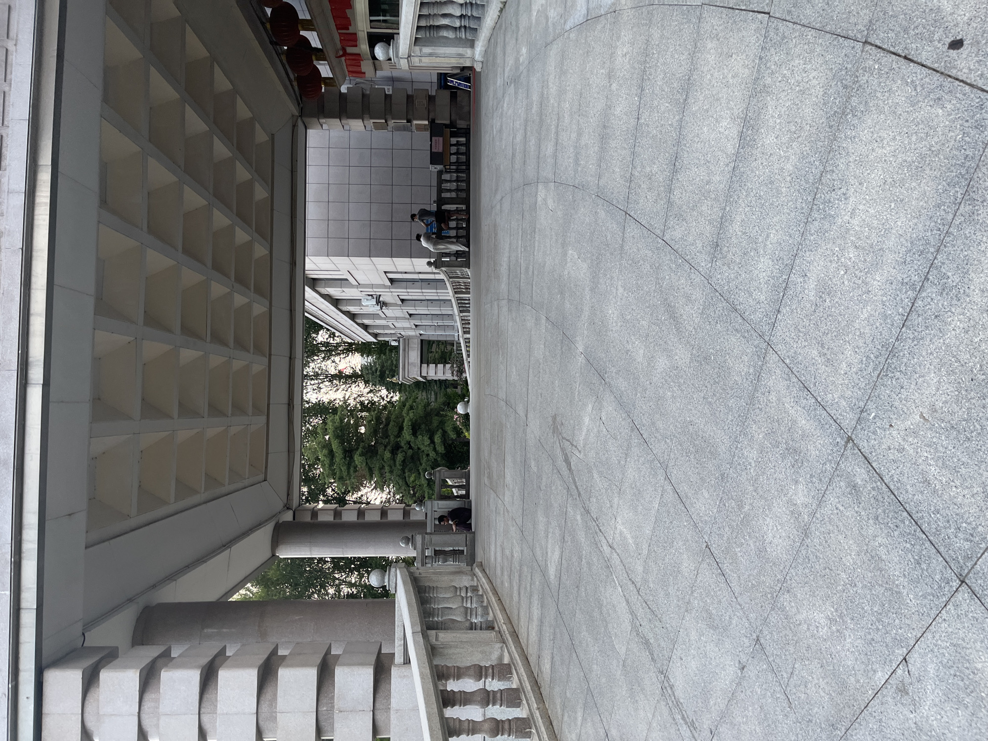 The image shows an outdoor walkway leading towards a building entrance. The walkway is paved with large grey granite tiles. On the left, there is a decorative stone railing supported by balusters. The building has a covered entrance with a large, stepped concrete ceiling structure. The building facade is made of grey tiled panels. Several people are visible in the background, some standing and talking, and one person sitting alone. Trees are visible behind the building. Red lanterns hang from the right side of the building's entrance, suggesting a festive or decorative element. The scene appears to be in a public area, possibly a government building or an institution, located in Changchun City, China.
