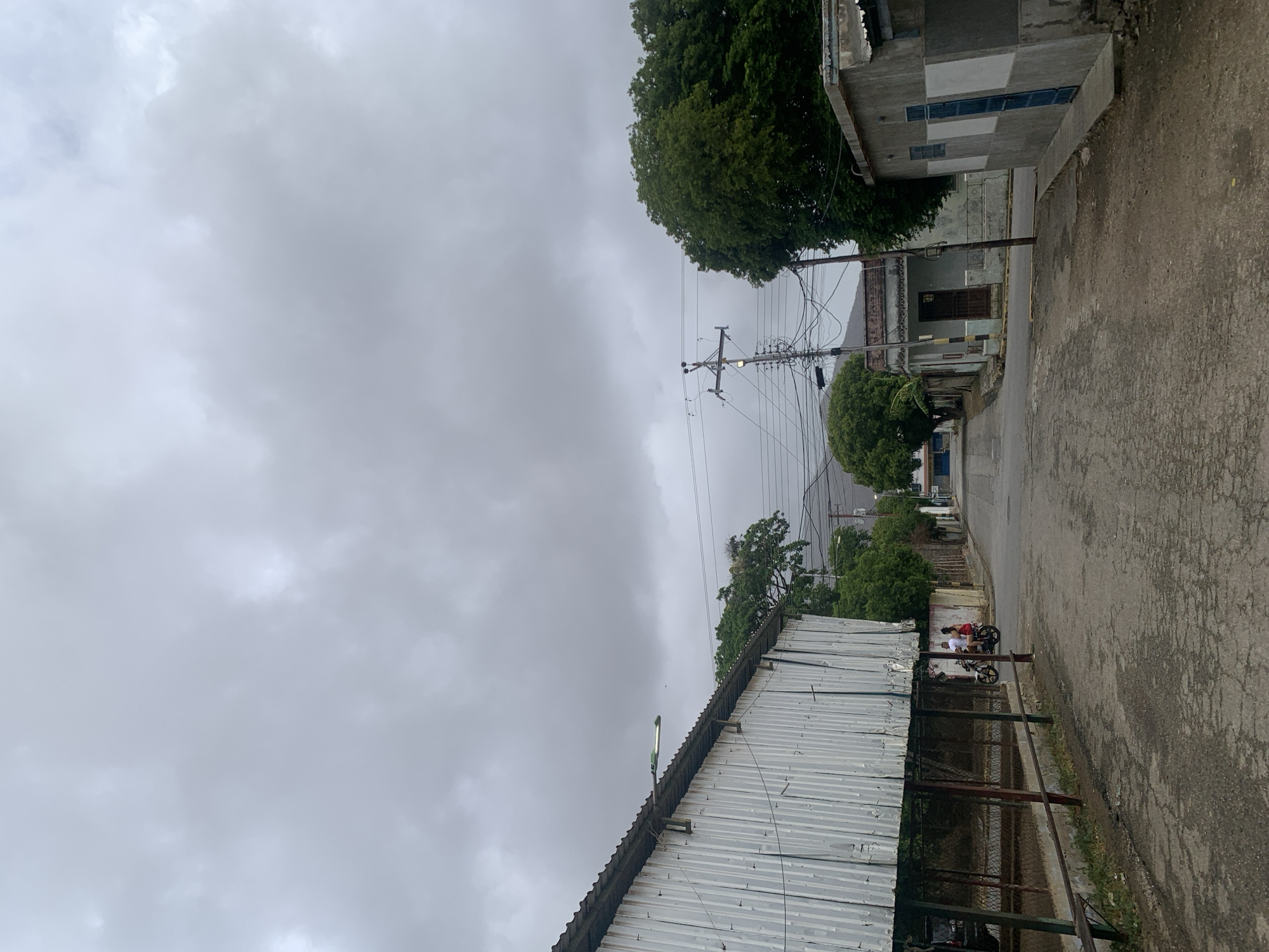 The image captures an overcast day on a street in Santa Ana, Venezuela, viewed from a slightly elevated perspective. The sky is dominated by thick, grey clouds, suggesting a cloudy or pre-rainy atmosphere.
In the foreground, a tall corrugated metal structure, possibly a fence or the side of a building, runs along the right side of the frame, with a modern, green-tinted street light attached to its roofline. A worn asphalt road, showing cracks and disrepair, stretches into the distance, bordered by a dirt and gravel shoulder on the right. Two people are visible on the side of the road, one sitting on and another standing next to a small red and black motorcycle.
Further along the street, on the left, stands a building with a dark entrance and a porch-like area. On the right, another building features concrete walls with a patterned design in various shades of grey and white, and blue rectangular elements, possibly vents or decorative tiles. Numerous vibrant green trees are scattered throughout the scene, providing natural foliage, particularly prominent in the mid-ground and background. A tall utility pole laden with a complex tangle of wires stands prominently in the middle distance, extending over the street. Another wooden pole is visible further down the road. The overall scene depicts a quiet, residential or semi-urban environment. No discernible text is visible in the image.