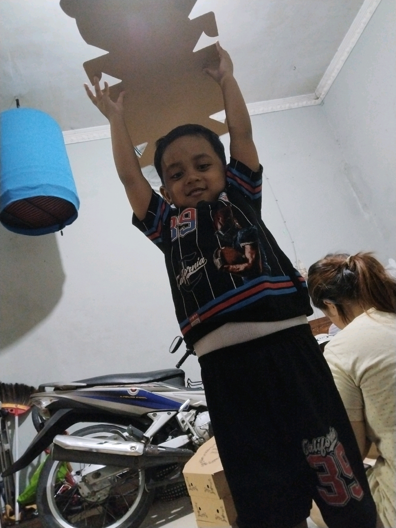 In a room, likely in Pepelegi, Indonesia, a young boy stands and holds a large, light brown cardboard cutout of what appears to be a bear shape above his head. He is wearing a black t-shirt with red and blue stripes on the sleeves and a graphic print, and black shorts with the number "39" in red and white. His arms are raised, holding the cardboard cutout. To the left of the boy, a blue cylindrical object, possibly a lampshade, hangs from the ceiling. In the background, a motorcycle is parked, and a woman with brown hair pulled back is visible, wearing a light-colored shirt and looking down. The room has white walls and a white ceiling with trim molding.