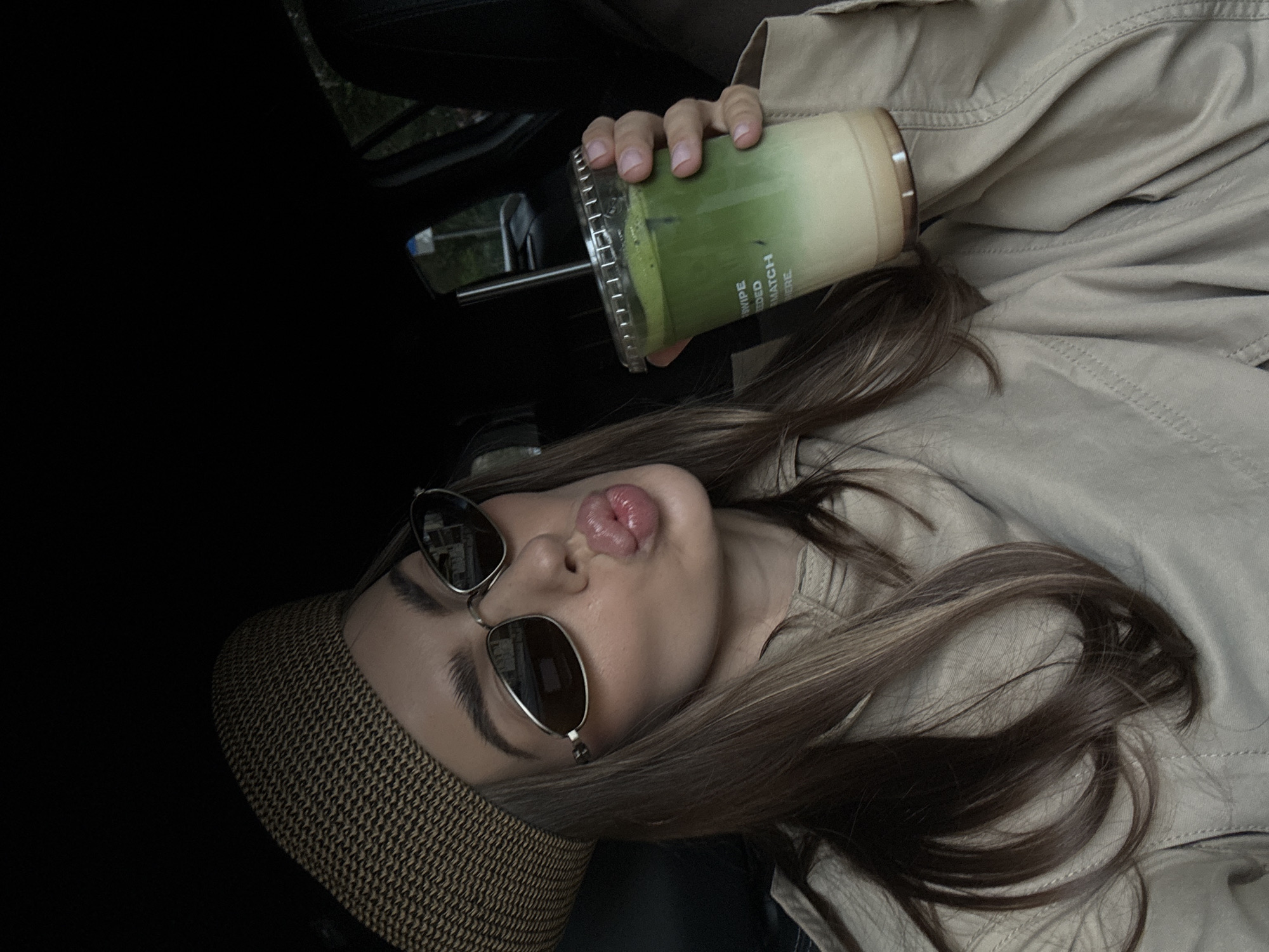 A young woman is lying in a car, wearing a woven hat and sunglasses. She is pouting her lips towards the camera and holding a large plastic cup of a matcha green drink in her right hand. She is wearing a light brown jacket, with long brown hair. It is daytime, and the location of the photograph may be Warsaw, Poland, but that cannot be determined from the image.