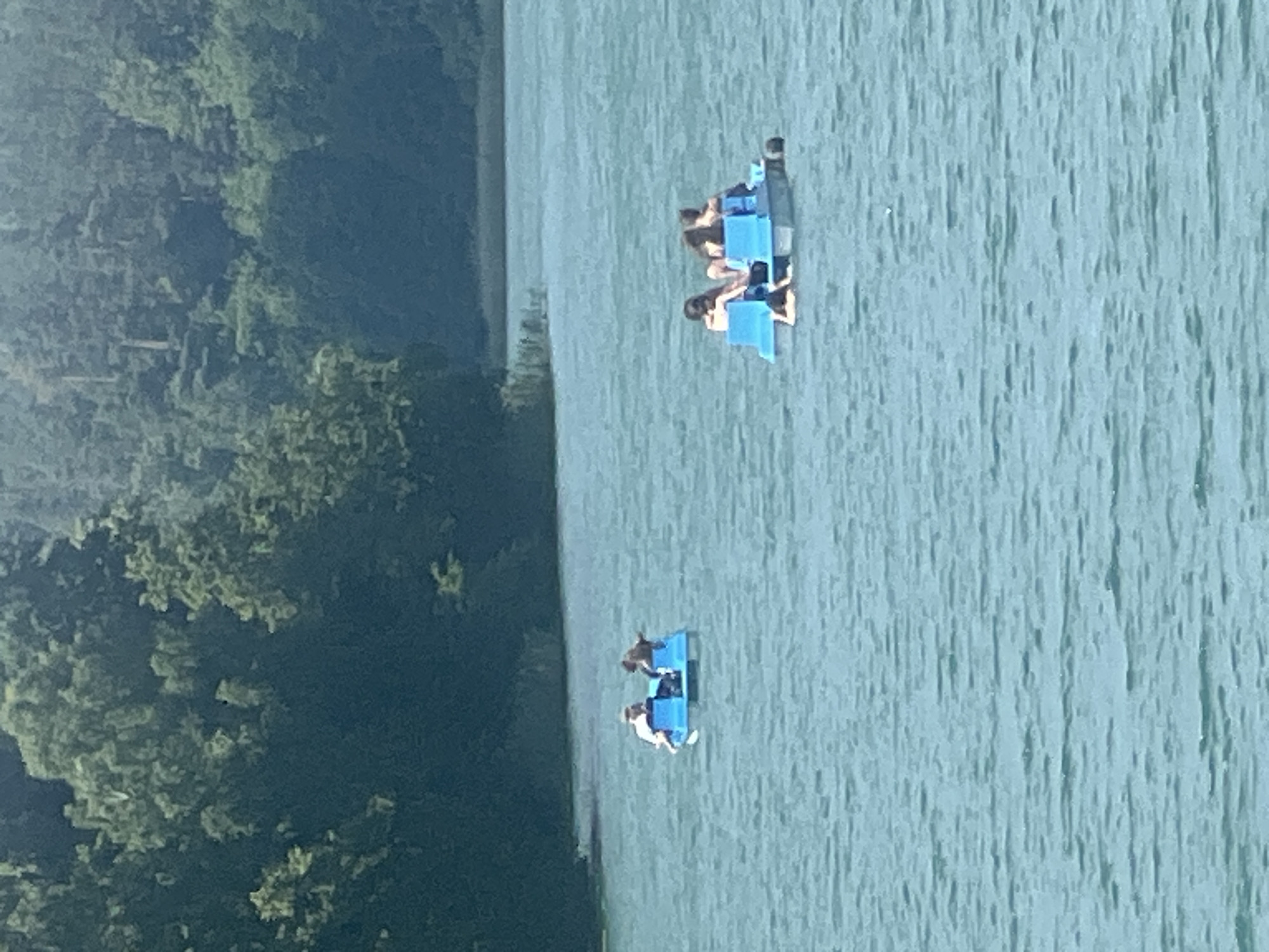 Two small blue pedal boats are on a large body of water. The boat in the foreground has two people on it, one wearing a white top and shorts and the other wearing dark clothing. The boat in the background has four people on it, all wearing dark clothing. The water is a light blue-green and appears calm. On the left side of the image, there is a densely wooded area with dark green foliage. The image is taken from a high angle, suggesting a vantage point above the water. The lighting suggests it is daytime, possibly late afternoon given the angle of the sun. The overall scene is one of recreation and leisure. The location is Vilnius, Lithuania.