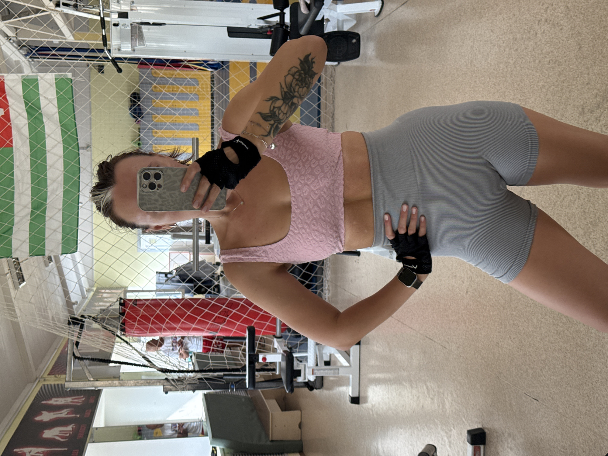 A woman is taking a selfie in a gym. She is wearing a pink crop top and grey workout shorts. She has a tattoo on her arm and is wearing black gloves and a smartwatch. The gym has various exercise machines and equipment, including a weight machine and a punching bag. The location is identifiable as Chaikovsky, Russia, based on the presence of the flag of Adygea visible in the mirror.