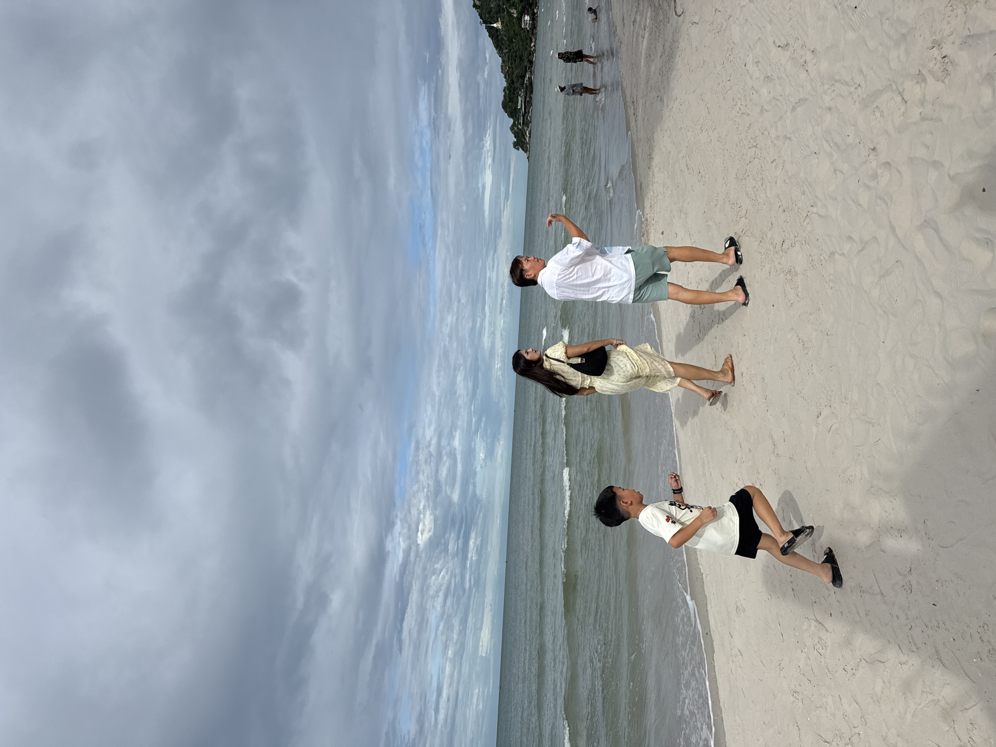 A family walks along a sandy beach in Hua Hin, Thailand, under a cloudy sky. The ocean stretches out to the horizon, with small waves lapping at the shore. In the foreground, a young boy in a white t-shirt and black shorts runs ahead of two adults. The woman is wearing a yellow dress and carrying a black purse, while the man is in a white shirt and light green shorts. In the background, other beachgoers can be seen wading in the water and walking along the shore. A verdant hill with a building on top is visible in the distance to the right.