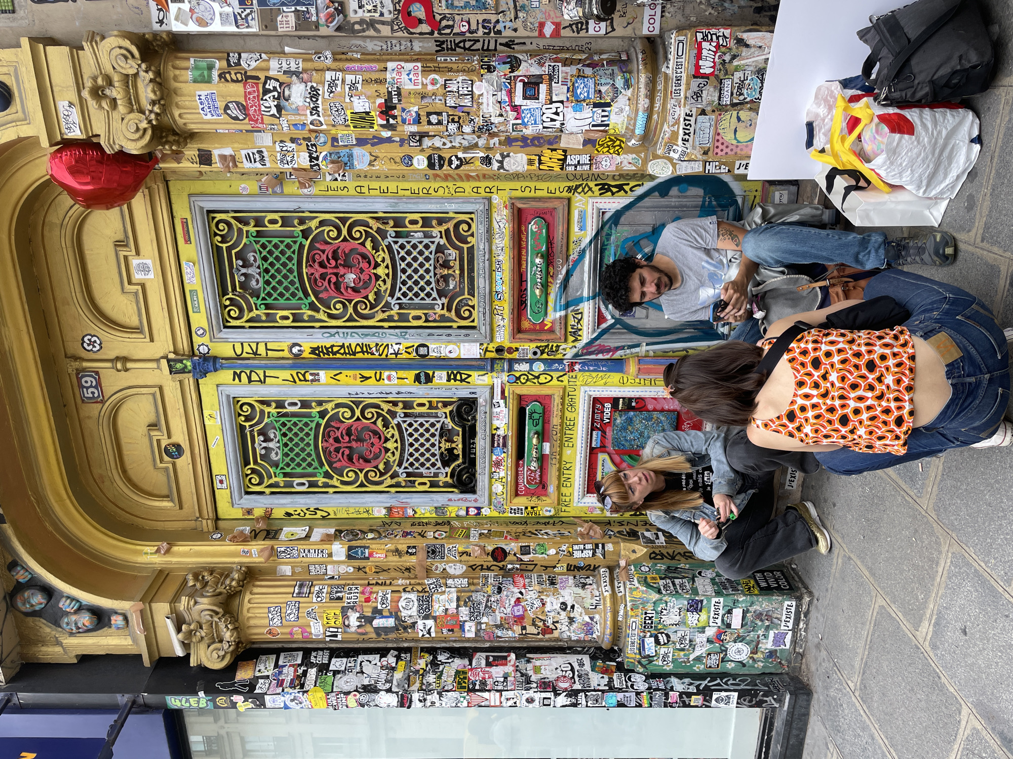 Three young adults are sitting on the sidewalk in front of a decorated doorway. The doorway is framed by golden pillars covered in stickers, and the double doors themselves are also adorned with various stickers and colorful ornate metalwork. A red heart-shaped balloon is tied to one of the pillars.
On the left, a woman with blonde hair is wearing a denim jacket and dark pants. She is looking towards the right, holding a small green object in her hand. In the center, a man with dark curly hair and a beard is wearing a grey t-shirt and blue jeans. He is looking down at his phone. On the right, a person wearing an orange and black patterned top and blue jeans is facing away from the camera, with their back to the viewer. They are also looking down.
There are several shopping bags and a dark duffel bag on the ground to the right of the people. The pavement is made of cobblestones. The scene suggests a casual outdoor gathering in a vibrant, sticker-covered urban setting. The presence of the number "59" above the doorway suggests a street address. While the exact location within Paris is not specified, the style of the doorway and the graffiti-like stickers point to a trendy, artistic neighborhood.