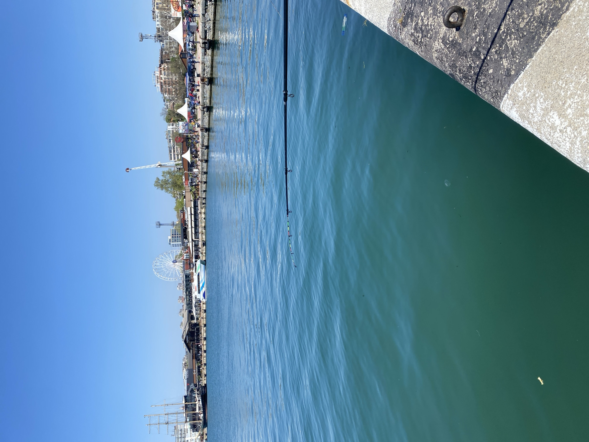 The image shows a view of the Varna harbor in Bulgaria, looking towards the city from a pier. Clear blue sky is visible in the background. In the water, there are fishing rods with floats attached to them, presumably being used for fishing. The harbor includes various attractions, such as a ferris wheel, tall ship, and several dining areas along the waterfront. On the pier, the edge is visible, suggesting the viewpoint is from the pier itself. The image is slightly angled counter-clockwise.