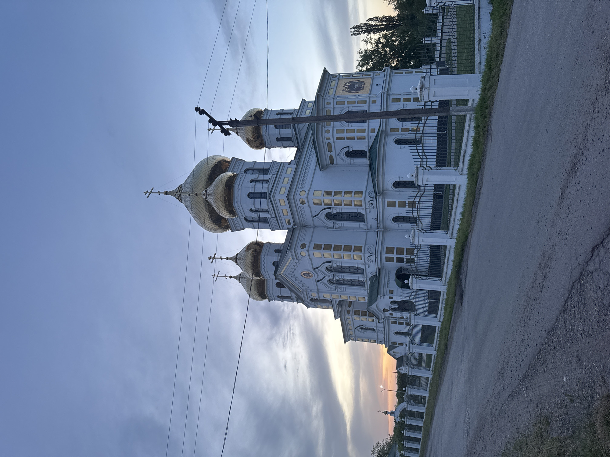 The image depicts the Holy Trinity Church in Novodonetskaya, Russia. The church is white with gold domes and crosses. It is positioned beside a road with a fence separating the church grounds from the road. Power lines run across the sky. The sky is a gradient of blue and orange, suggesting either sunrise or sunset.