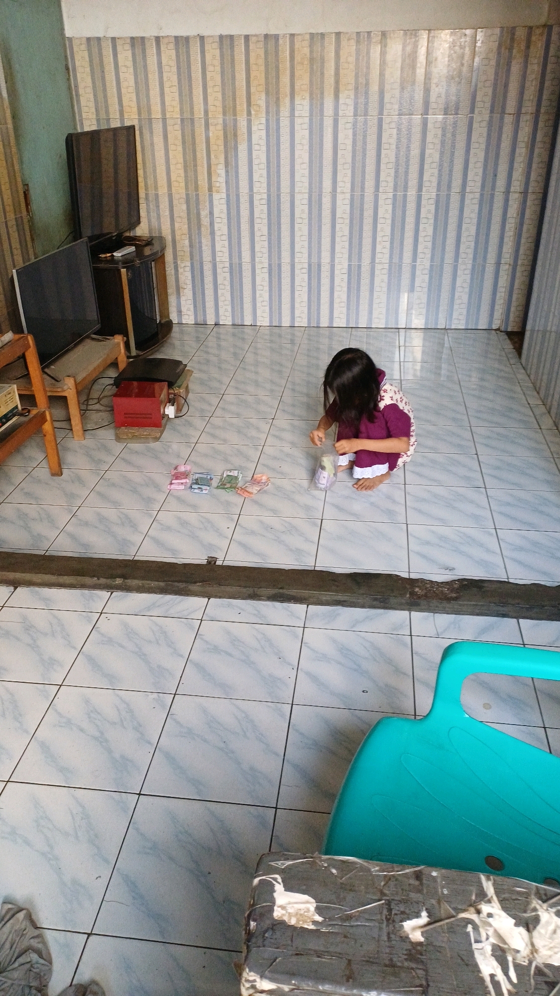 A young girl with dark hair, wearing a purple and white patterned dress, is kneeling on a tiled floor in what appears to be an outdoor covered area. She is holding a clear plastic bag and is engaged in an activity with several banknotes laid out in front of her. The banknotes are of various colors and denominations, suggesting a financial transaction or counting activity.
The environment is simple and functional, with a tiled floor and a wall in the background covered in a striped wallpaper pattern. To the left of the girl, there is a wooden chair and a television set on a stand, along with a red electronic device. In the foreground, a bright turquoise plastic object and a dark, taped-up rectangular object are visible.
The scene is consistent with a residential setting in Gunung Putri, Indonesia, a region known for its mix of rural and developing urban characteristics. The overall impression is one of a candid snapshot of everyday life.