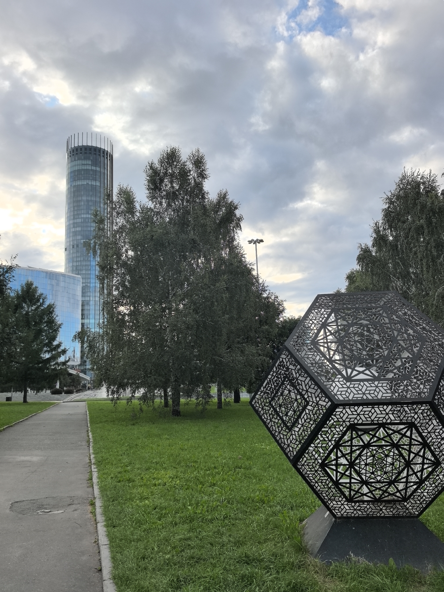 In Yekaterinburg, Russia, a modern city park scene unfolds under a cloudy sky. In the background, the striking Iset Tower, a cylindrical skyscraper with a distinctive crown of vertical elements, dominates the skyline. Adjacent to it stands a modern building with a blue glass facade. The foreground is occupied by a pathway running alongside a lush green lawn. To the right of the pathway, a large, geometric sculpture with intricate black lattice-work, resembling a geodesic dome, is prominently displayed. Several trees, including birches, are scattered throughout the park. A lamppost is visible in the mid-ground, and in the distance, the outline of a fountain can be discerned.