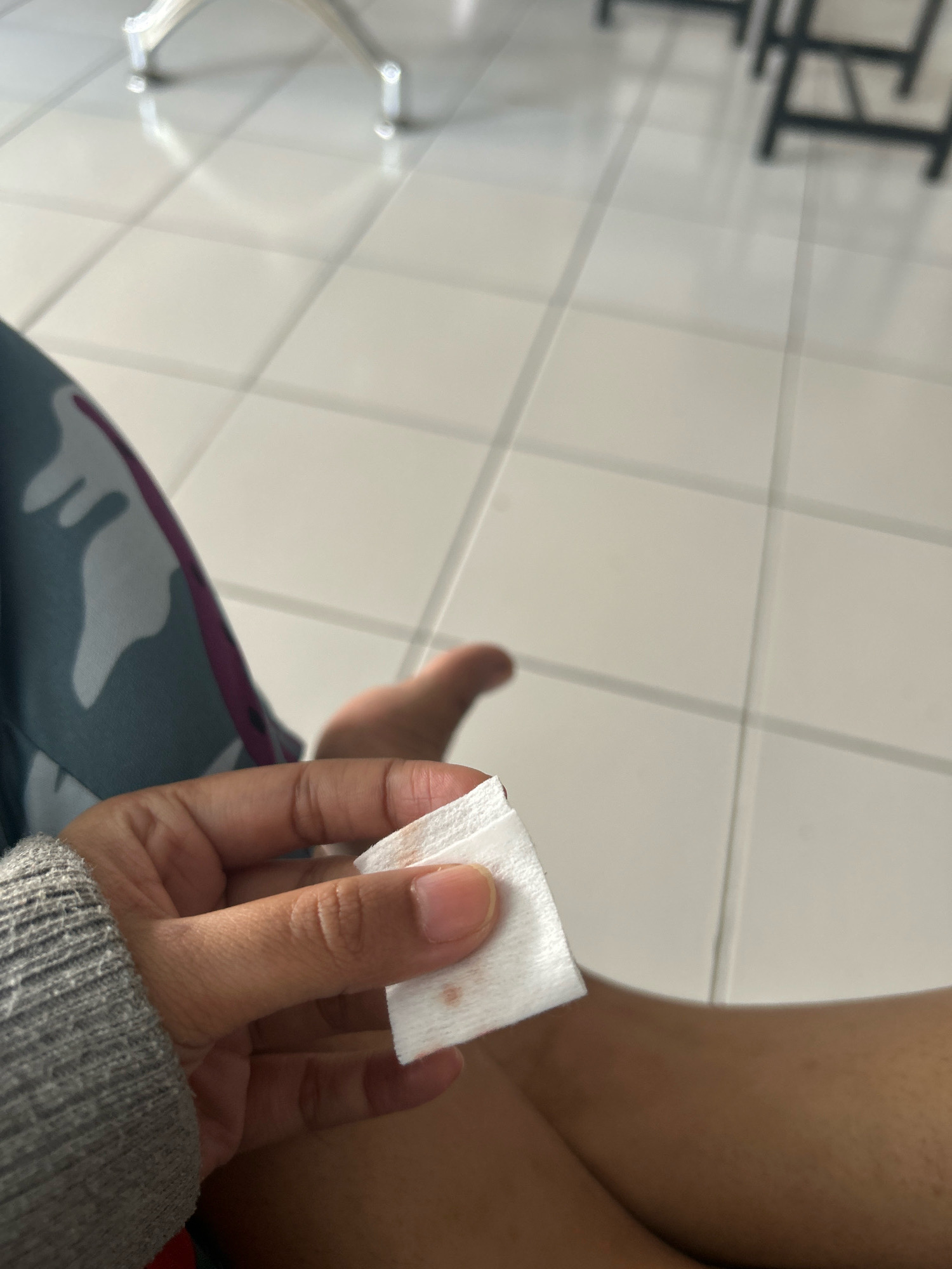 A person's hand is holding a small white cotton pad with a faint reddish stain on it. The hand is resting on the person's lap, which is covered by clothing with a camouflage pattern. The background shows a tiled floor and out-of-focus furniture, suggesting an indoor setting. The scene does not provide enough information to determine the specific location within Sukasetia, Indonesia.