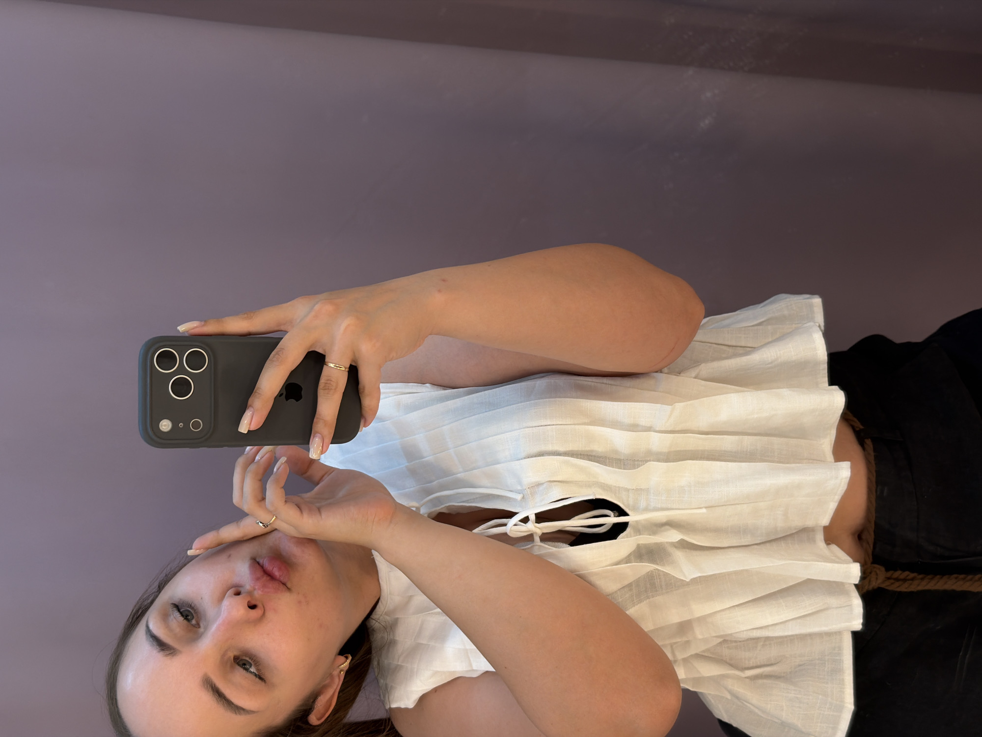 A woman is holding a gray iPhone in front of her face, taking a selfie. She is wearing a white blouse with a tie front and black bottoms. She has a gold ring on one finger. The background is a light purple wall. This image does not contain information that would indicate the location is in the city of Chaikovsky, Russia.