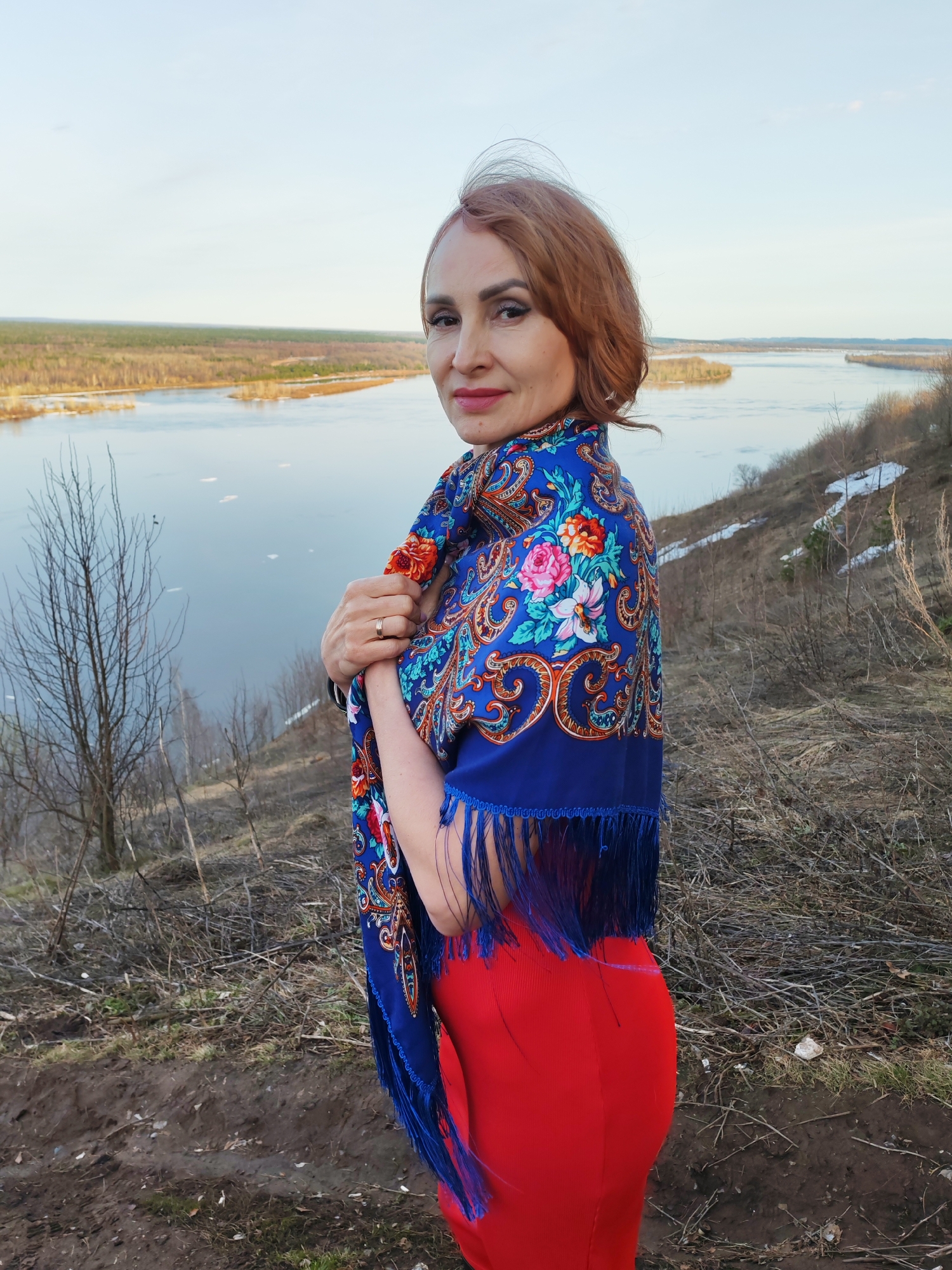 A woman stands on a hillside overlooking a river. She is dressed in a red garment and a blue shawl with floral patterns and a fringed edge. The river has ice floating on it. The hillside is covered in dry vegetation. The location is likely Sarapul, Russia, due to the geography and the woman's traditional Russian shawl.