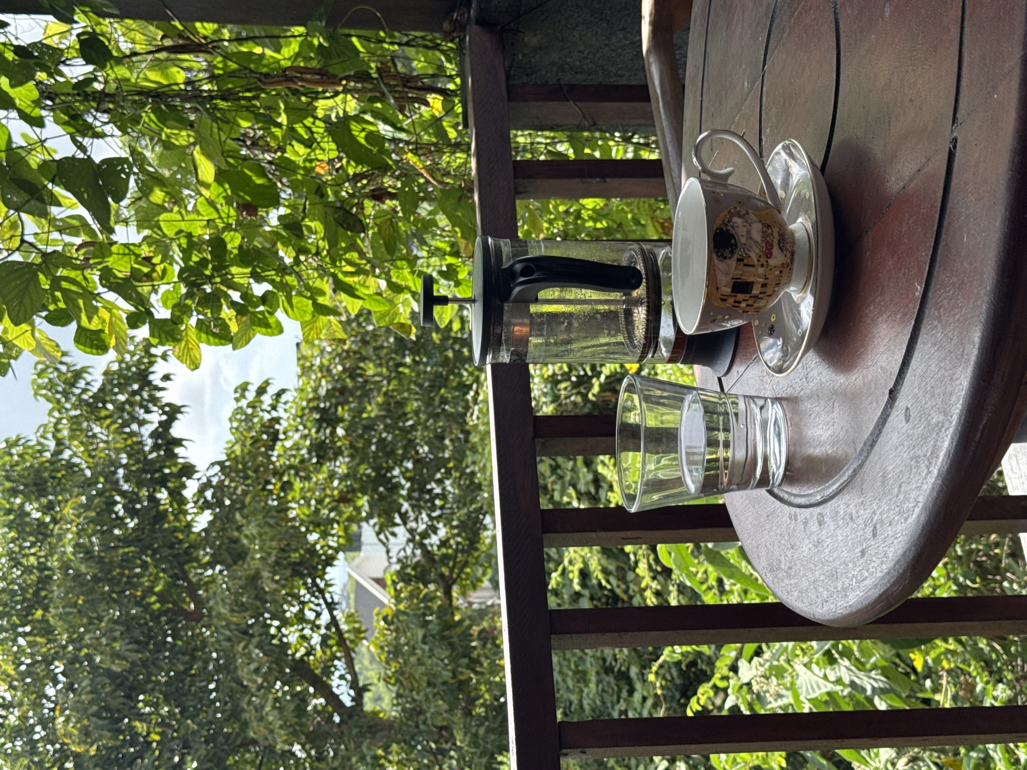 On a partly sunny day, a table on a terrace or balcony overlooking lush green trees and foliage, possibly in Kuta Selatan, Indonesia, hosts several items related to hot beverages.
The table, made of dark stained wood, is round and has a raised edge. Resting on it are a clear glass French press coffee maker, a glass of water, and a decorative teacup with an accompanying saucer. The teacup features artwork reminiscent of Gustav Klimt’s “The Kiss.”
The setting suggests a relaxed, outdoor leisure moment. The foliage obscures a full view, hinting at a residential neighborhood. A wooden fence or balustrade is visible in the background, supporting the terrace or balcony. The overall image presents a tranquil and aesthetically pleasing scene.