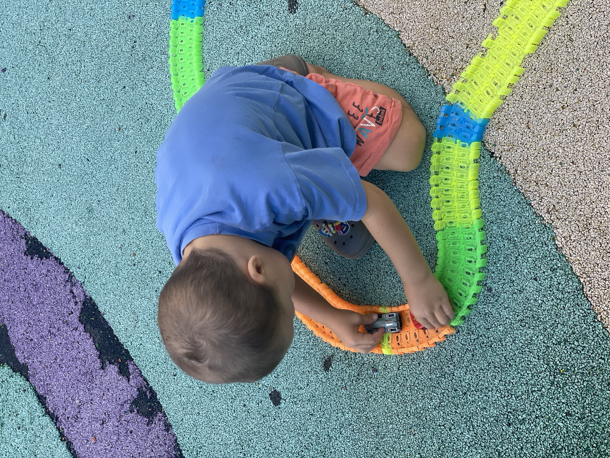 A young boy kneels on a multi-colored rubberized playground surface, assembling a bright orange and green glow-in-the-dark race track. He is wearing a blue t-shirt and peach-colored shorts with white lettering that reads "WAVES" and a small logo. The boy is holding a grey toy car and appears to be placing it on the track. The playground surface is a mix of turquoise, purple, and light brown speckled areas. The image was taken at the "Sochi Park" amusement park in Sochi, Russia, given the distinctively designed rubberized playground surfacing.