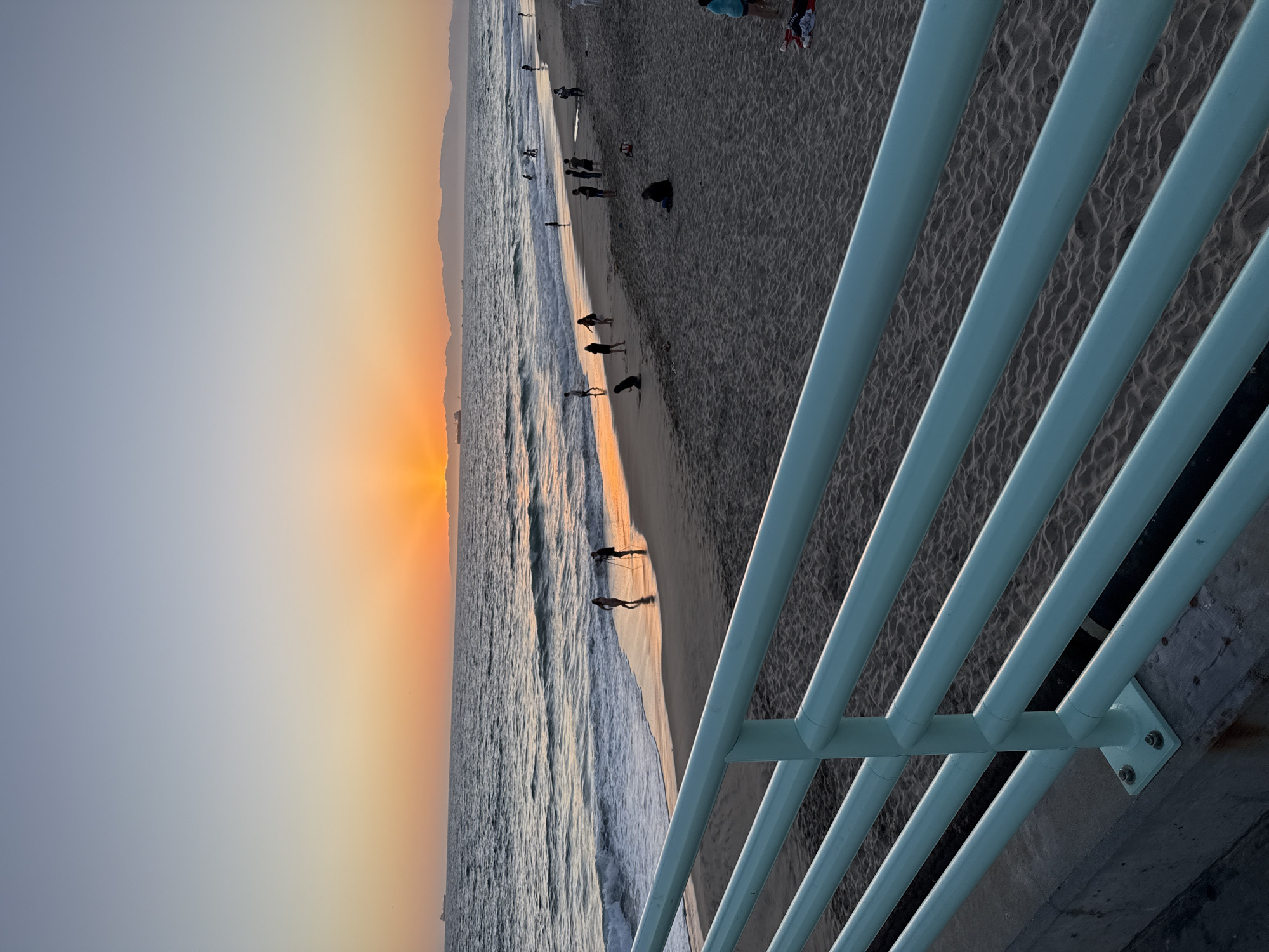 This image captures a sunset scene at Manhattan Beach, California, USA. The foreground is dominated by a pale blue railing that runs diagonally across the frame. Beyond the railing, a sandy beach stretches out, dotted with several people. Some individuals are walking along the shoreline, while others are sitting or standing on the sand. The ocean is visible in the midground, with gentle waves lapping at the shore and reflecting the warm hues of the sunset. Several ships are visible on the horizon, and a mountain range can be seen in the far distance. The sky transitions from a pale blue at the top of the frame to a vibrant orange and yellow near the horizon where the sun is setting. The overall atmosphere is serene and tranquil.