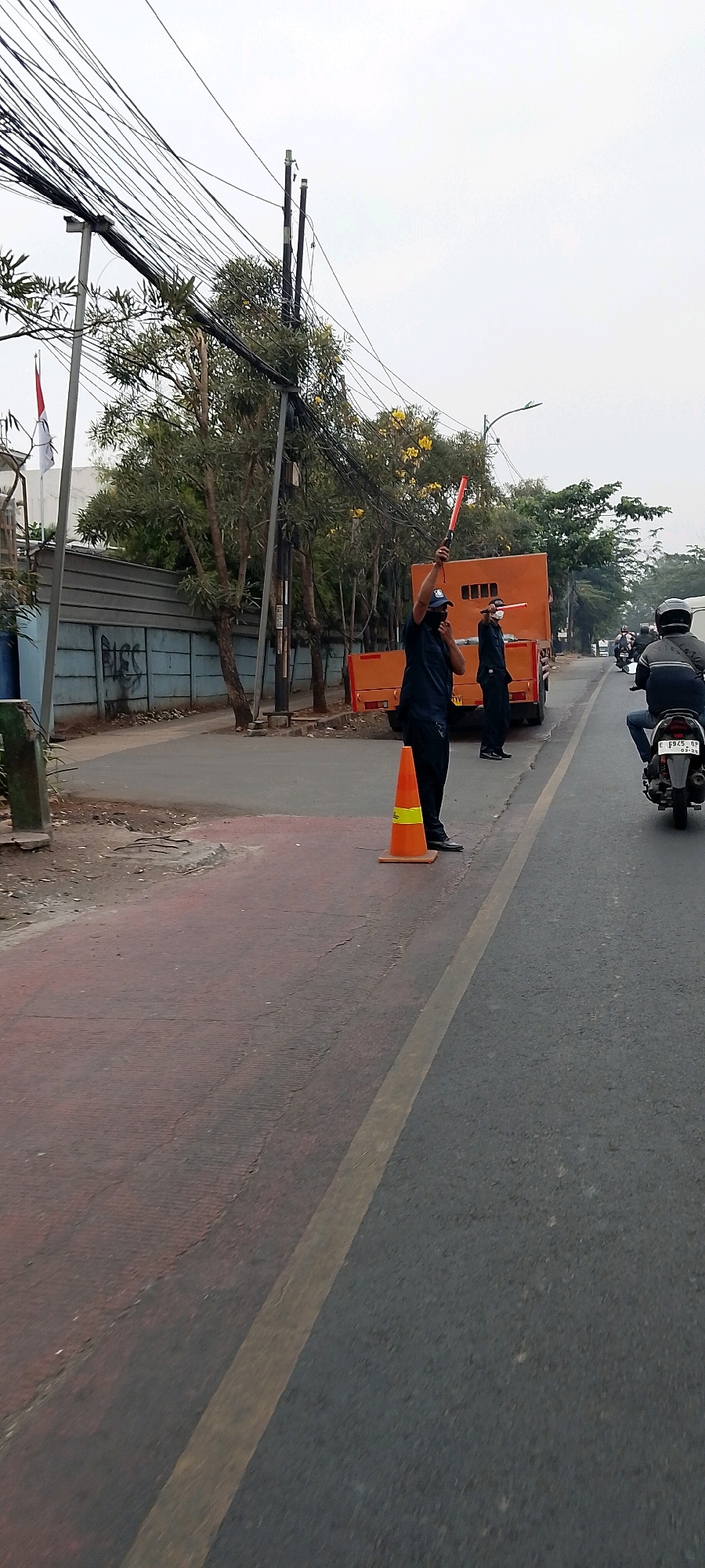 In Bekasi, Indonesia, on a street with overhead power lines and a grey fence with graffiti, two traffic directors wearing dark uniforms are directing traffic. One director, positioned near an orange traffic cone, holds a red signaling baton above his head. The second director stands beside an orange truck, also holding a red signaling baton, held horizontally. A motorbike rider in a black helmet is visible in the right lane of the road. The road surface is asphalt, with a reddish-colored lane to the left, separated by a yellow line. Trees with yellow flowers are present in the background, along with a small flag of Indonesia on a pole. The sky is overcast.